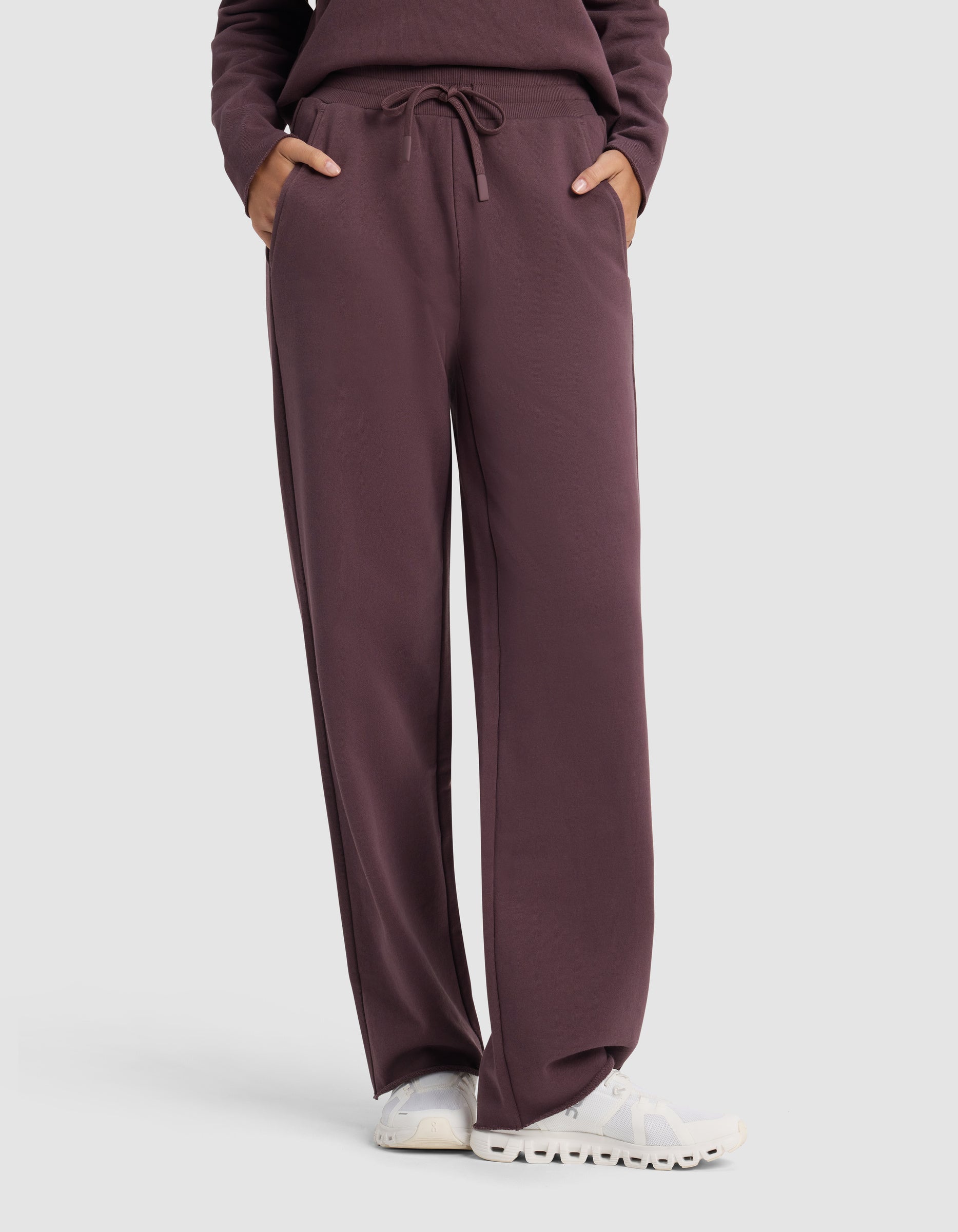Model wearing Cozy Earth Chelsea Pant in dark maroon with hands in pockets, paired with a matching top and white sneakers, standing against a plain light background.|Color:Fig