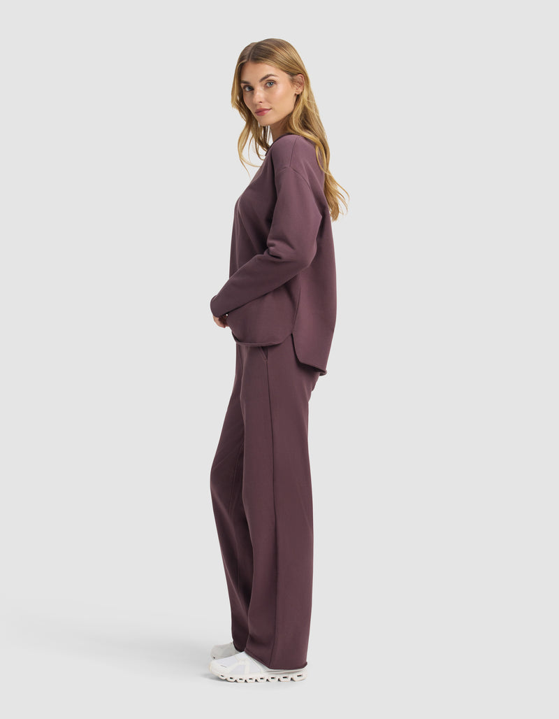 A woman stands in profile wearing the Cozy Earth Women's Chelsea Set—loose-fitting, long-sleeve dark mauve sweatshirt and matching pants—with white sneakers, set against a plain light background.