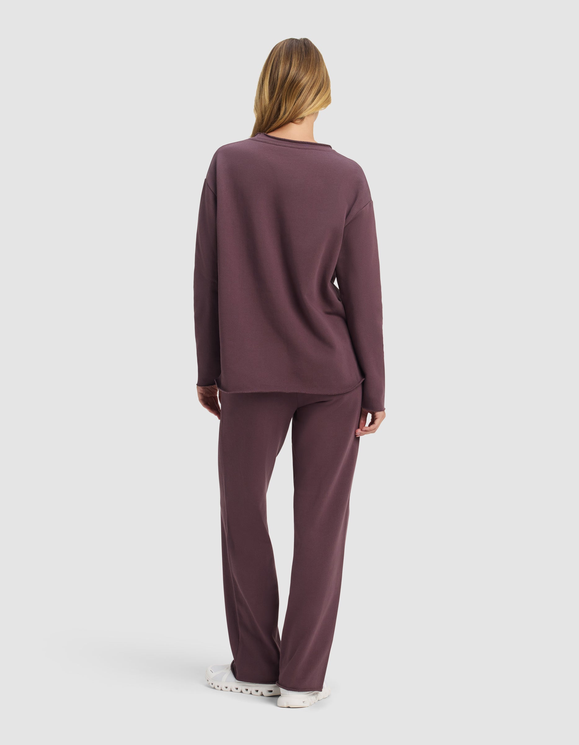 A woman with long hair, seen from behind, wears the Cozy Earth Women's Chelsea Set—a loose-fitting, long-sleeve top and matching pants in muted purple—paired with white shoes against a plain white background.|Color:Fig
