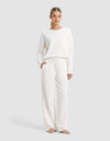 A woman stands barefoot against a light gray background, wearing the Cozy Earth Women's Chelsea Set: a loose, long-sleeve white top paired with matching wide-leg drawstring pants.|Color:Bone