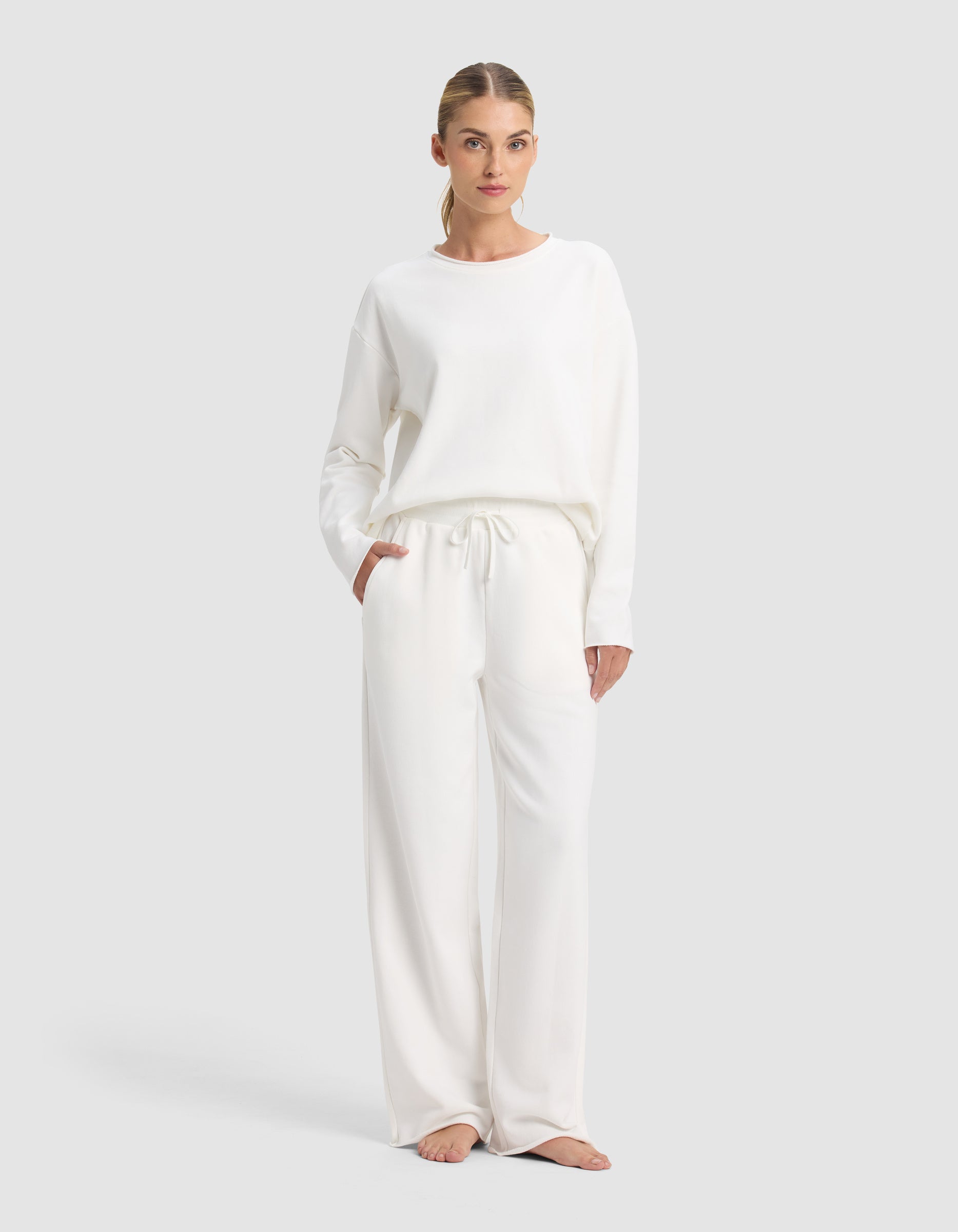 A woman stands barefoot against a light gray background, wearing the Cozy Earth Women's Chelsea Set: a loose, long-sleeve white top paired with matching wide-leg drawstring pants.|Color:Bone