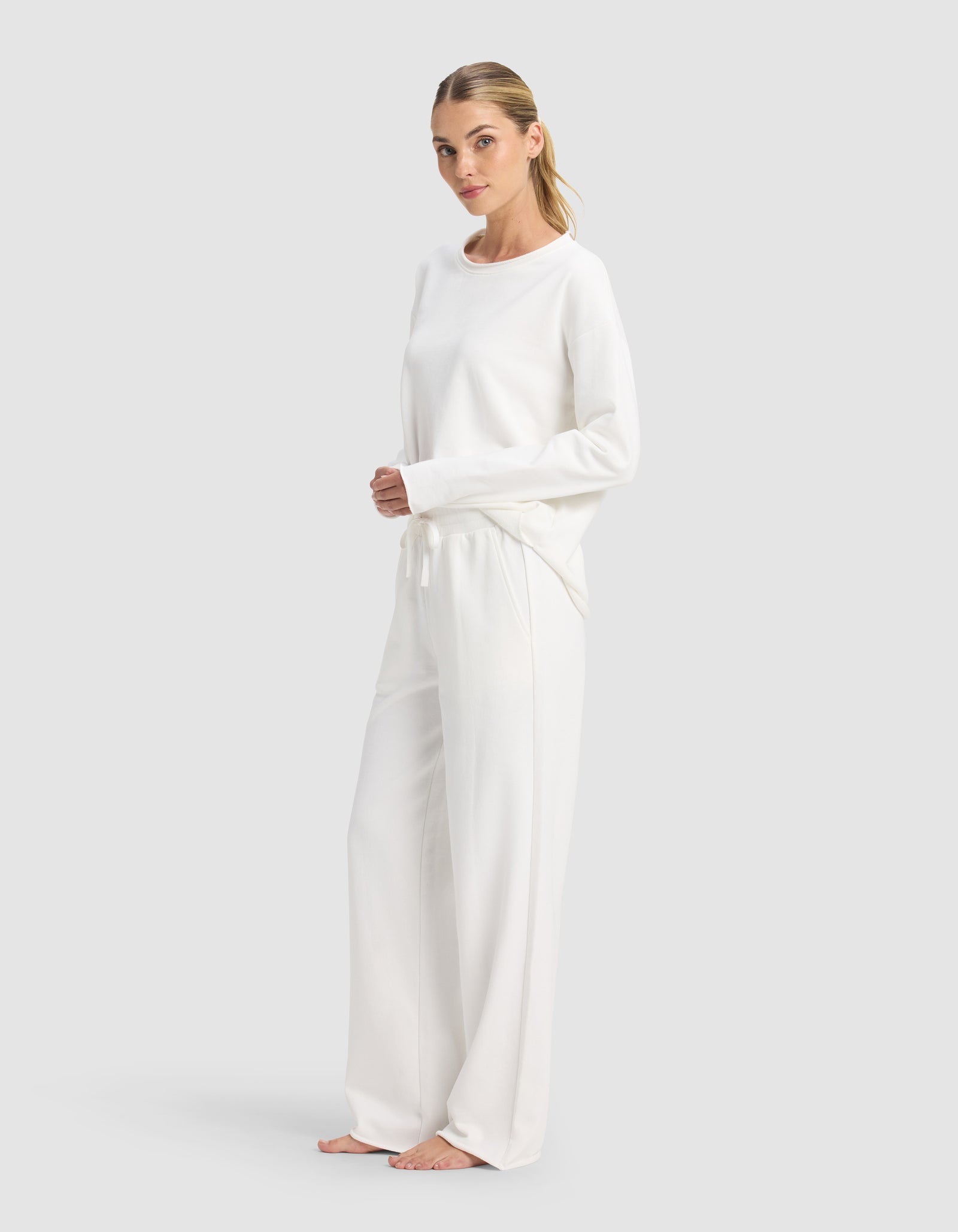 A woman with light hair pulled back stands barefoot against a plain backdrop, calmly facing the camera in the Cozy Earth Women's Chelsea Set—a loose long-sleeve top and wide-leg lounge pants, both in white.