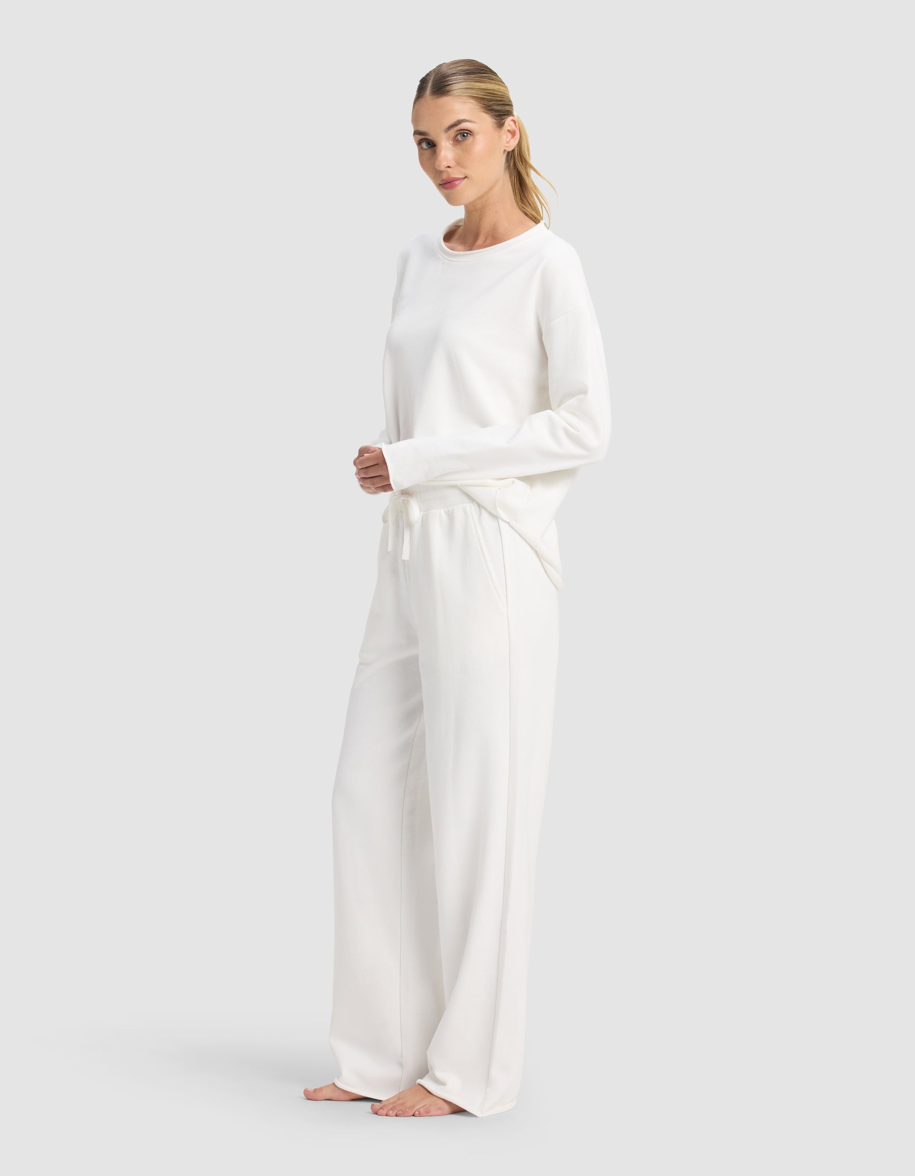 A woman with light hair pulled back stands barefoot against a plain backdrop, calmly facing the camera in the Cozy Earth Women's Chelsea Set—a loose long-sleeve top and wide-leg lounge pants, both in white.|Color:Bone