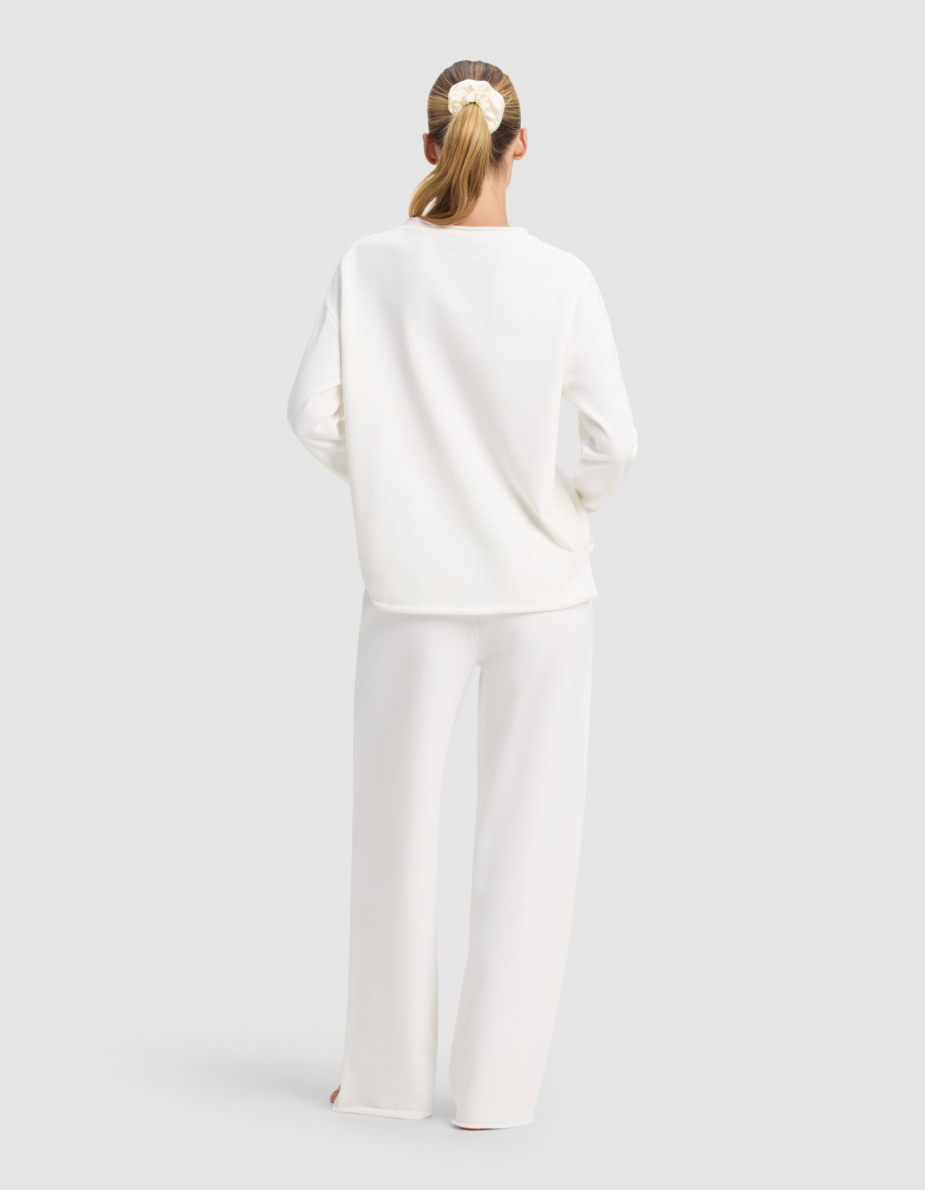 A woman with a ponytail and scrunchie stands with her back to the camera, wearing the Cozy Earth Women’s Chelsea Set—loose-fitting white long-sleeve top and wide-leg pants—against a plain light gray background.|Color:Bone
