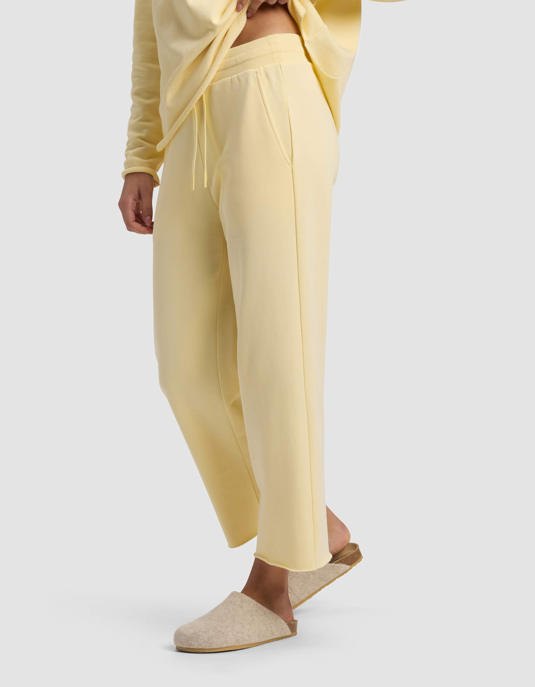 A model wears Cozy Earth's Women's Chelsea Cropped Pant in pale yellow with a matching top, one foot slightly raised to show beige slippers. The background is plain light gray, and the model’s face is not visible.