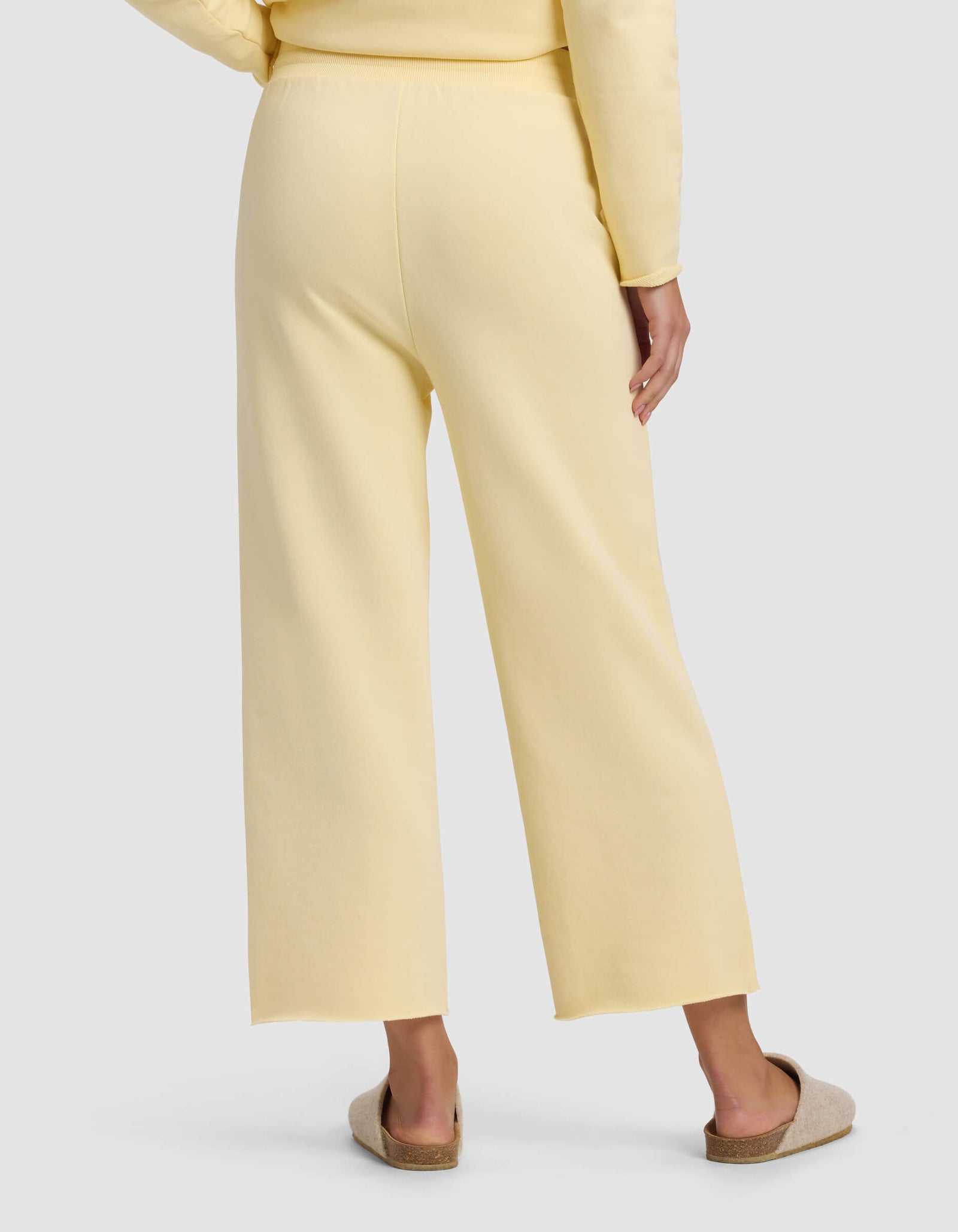 A person is seen from the back wearing Cozy Earth's Women's Chelsea Cropped Pant in light yellow with a matching top, standing on a white surface and beige slip-on slippers.