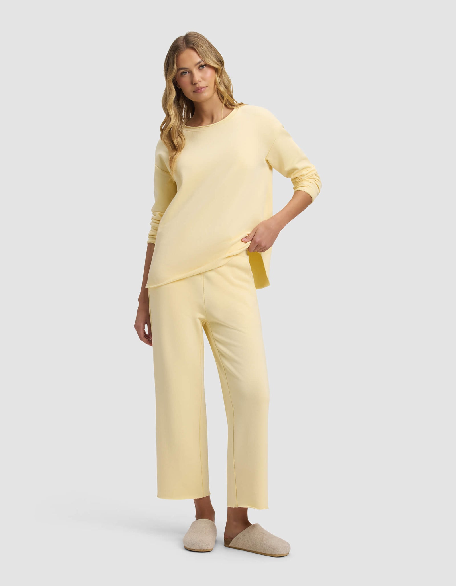 A woman with long blonde hair wears Cozy Earth's Women's Chelsea Cropped Pant in pale yellow with a matching loose top and beige slippers, standing against a plain light gray background.