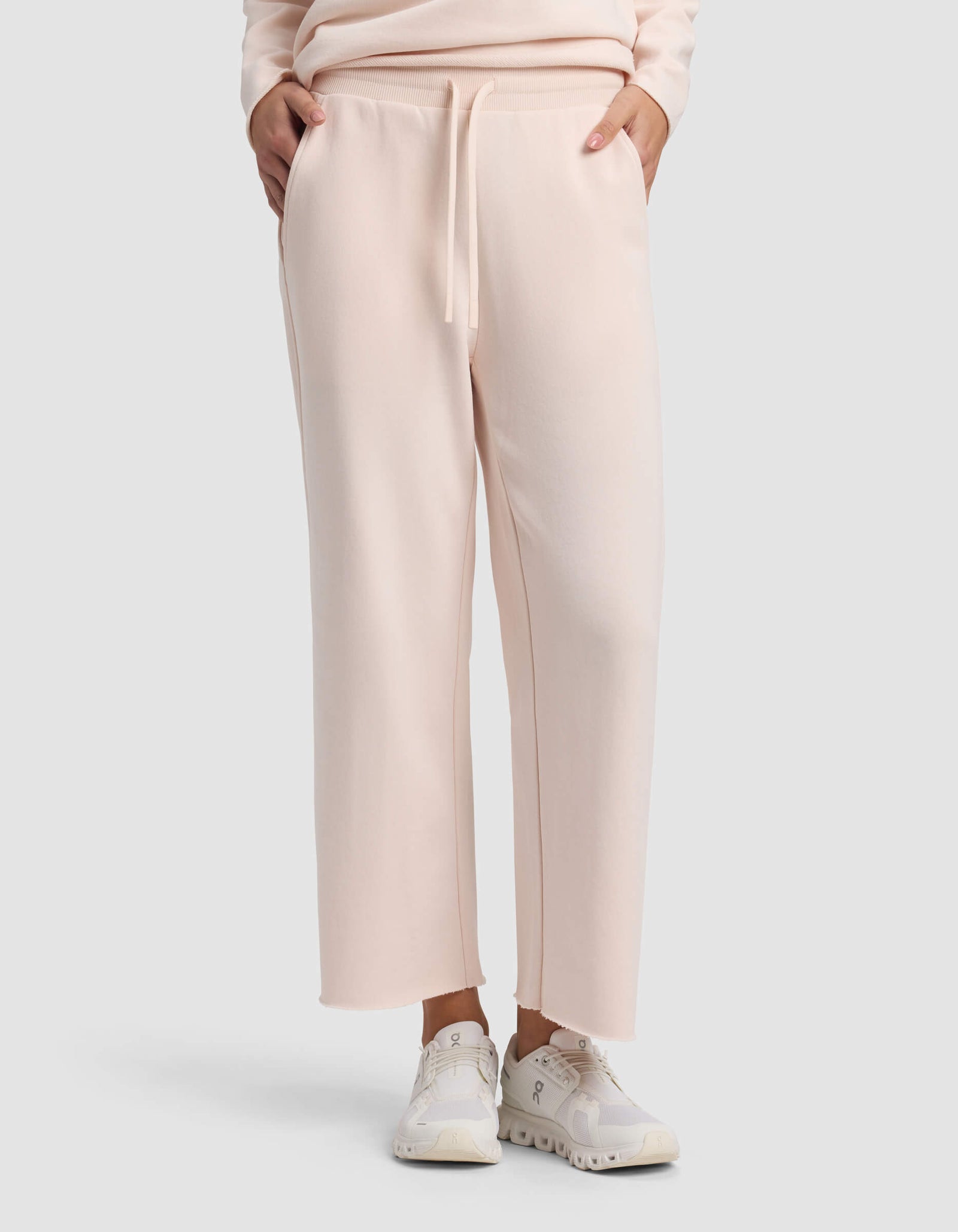 A person wears Cozy Earth Women's Chelsea Cropped Pant in light pink, styled with white sneakers and hands in pockets, against a plain light background.