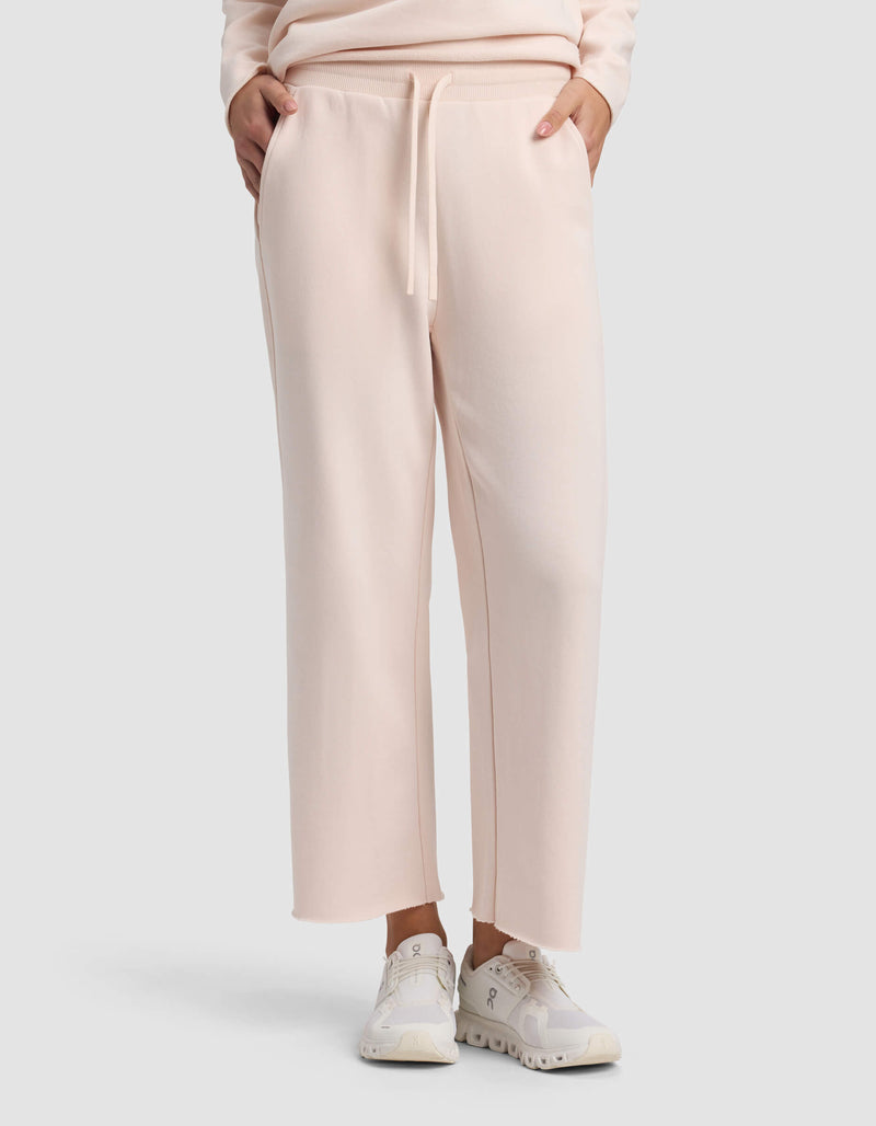 A person wears Cozy Earth Women's Chelsea Cropped Pant in light pink, styled with white sneakers and hands in pockets, against a plain light background.