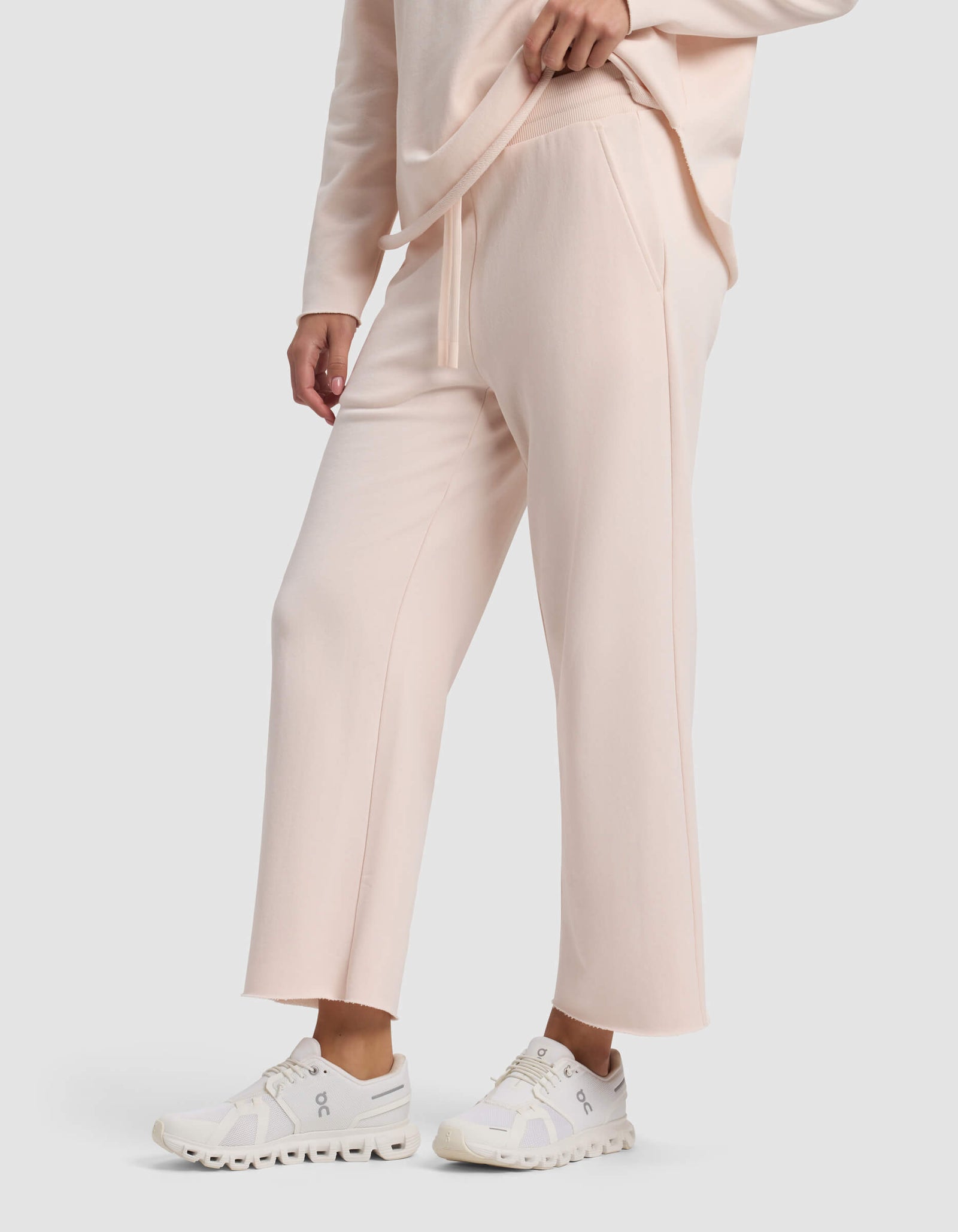 A person wears Cozy Earth's Women's Chelsea Cropped Pant in light pink with a matching sweatshirt and white athletic sneakers, standing with one hand in their pocket against a plain light gray background.