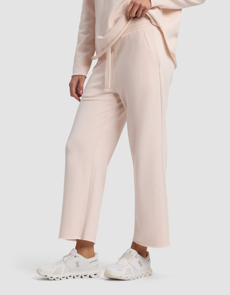 A person wears Cozy Earth's Women's Chelsea Cropped Pant in light pink with a matching sweatshirt and white athletic sneakers, standing with one hand in their pocket against a plain light gray background.