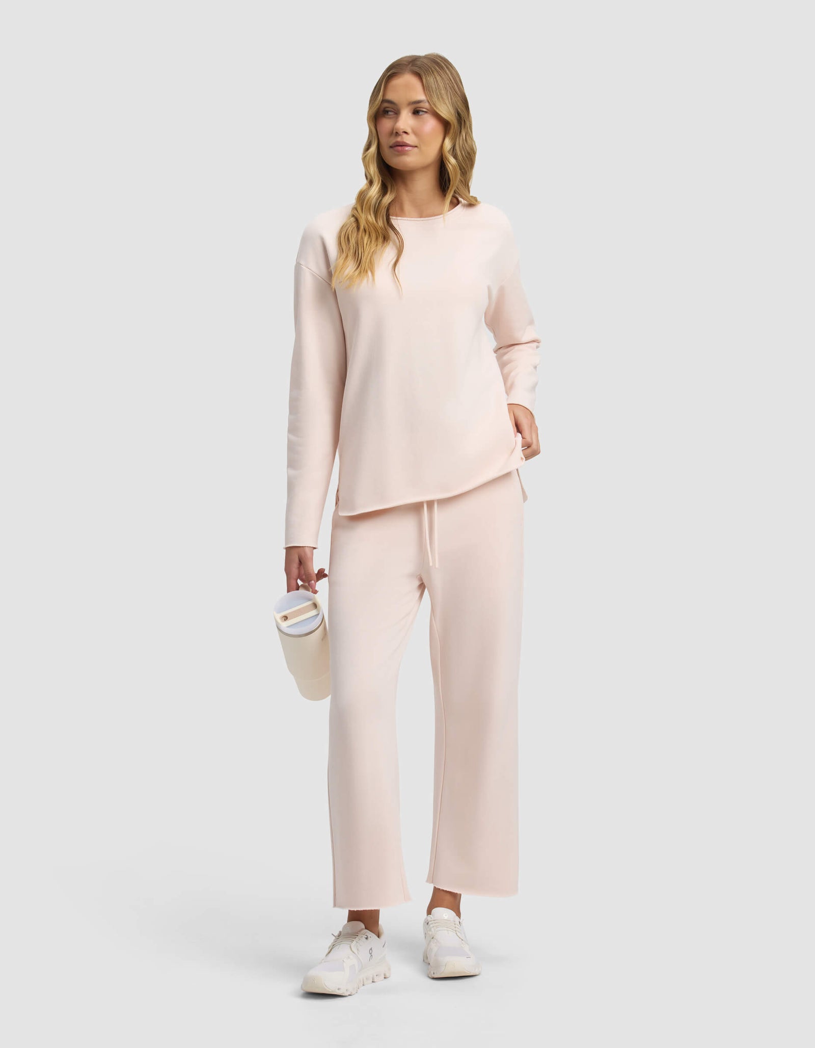 A woman with long wavy blonde hair wears Cozy Earth's Women's Chelsea Cropped Pant and a light pink long-sleeve top, paired with white sneakers, holding a white tumbler against a plain light gray background.