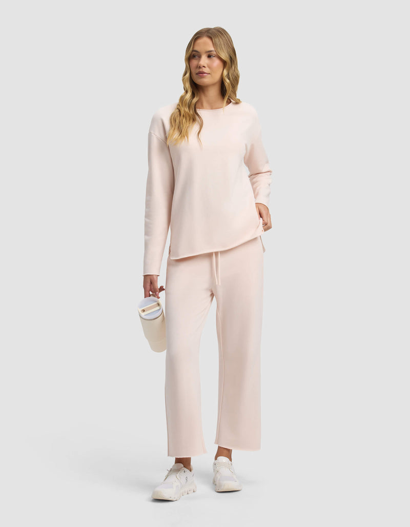 A woman with long wavy blonde hair wears Cozy Earth's Women's Chelsea Cropped Pant and a light pink long-sleeve top, paired with white sneakers, holding a white tumbler against a plain light gray background.