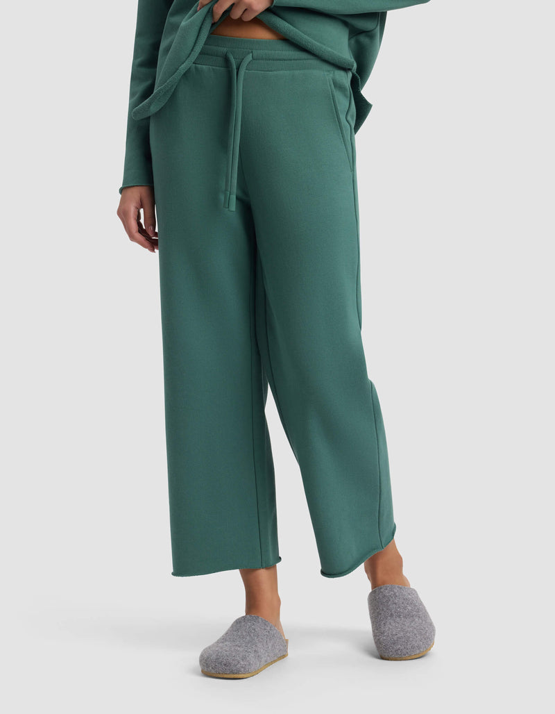 A person wears the Cozy Earth Women's Chelsea Cropped Pant in green with a drawstring, a matching top partly visible, and gray slippers, standing against a plain light gray background.