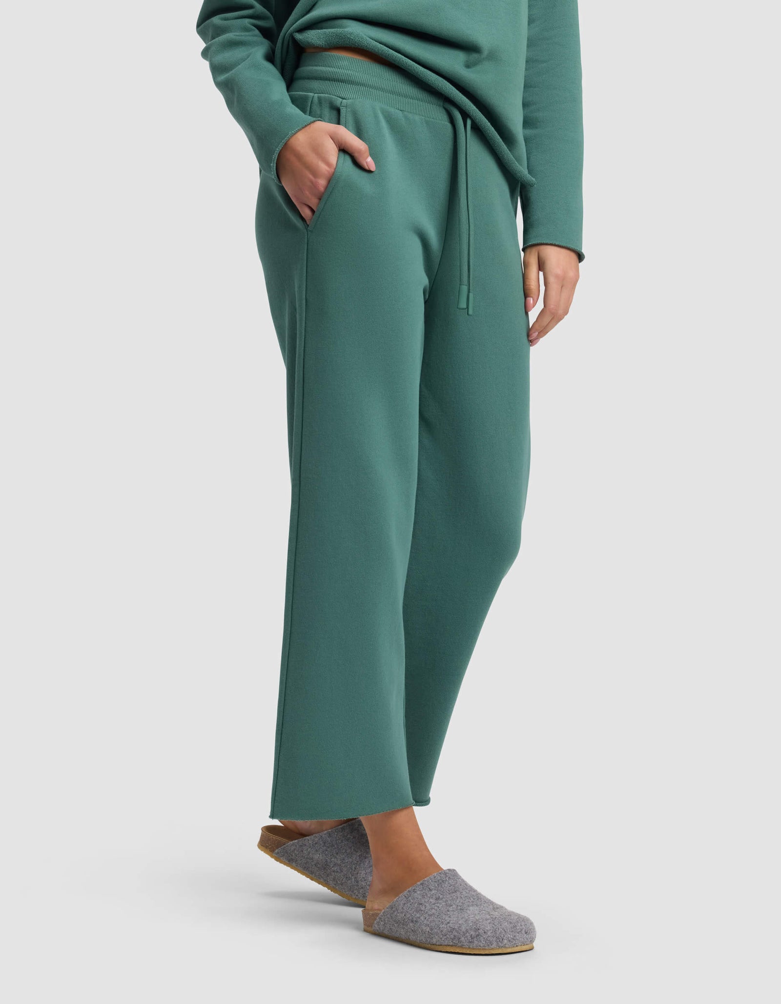 A person stands against a plain background with one hand in their pocket, wearing Cozy Earth's Women's Chelsea Cropped Pant in green, paired with a matching green sweatshirt and gray felt slippers.