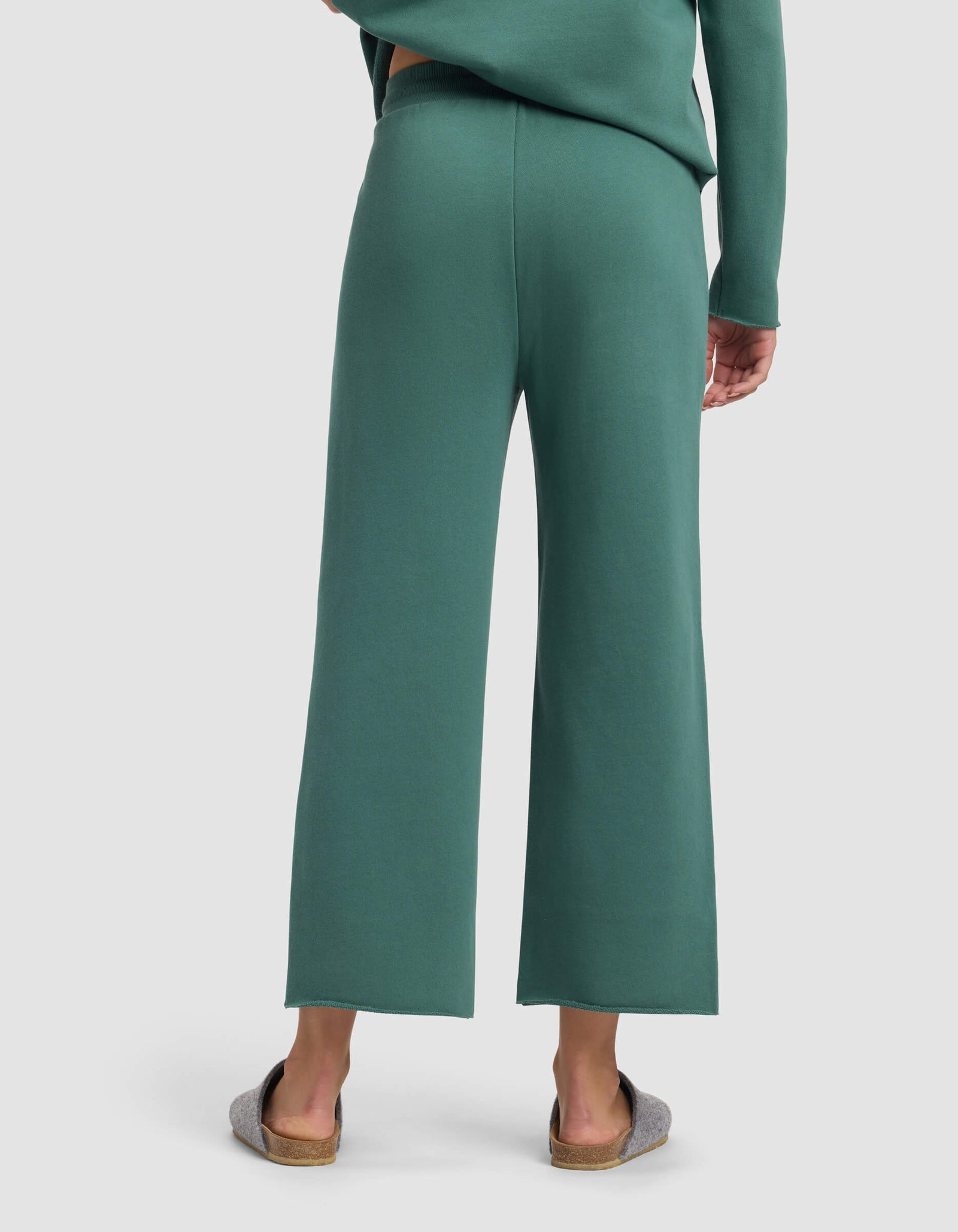 A person is shown from the back wearing Cozy Earth’s Women's Chelsea Cropped Pant in green with a matching top and gray slippers, standing against a plain light background.