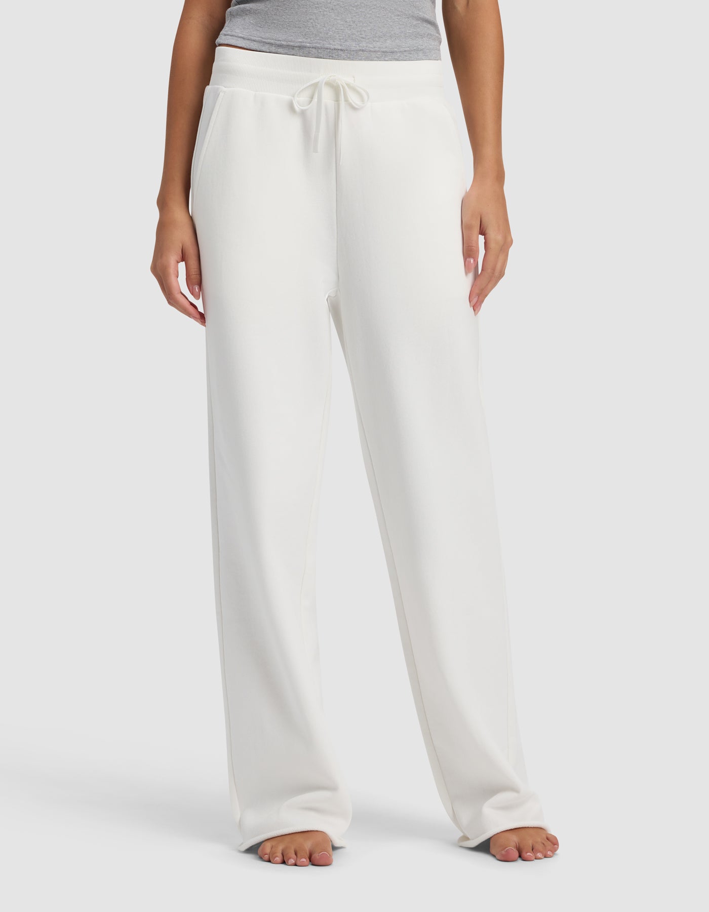 A woman models the Cozy Earth Women's Chelsea Pant, a loose-fitting, white drawstring lounge pant. She stands barefoot against a plain light background, with only her lower half visible.