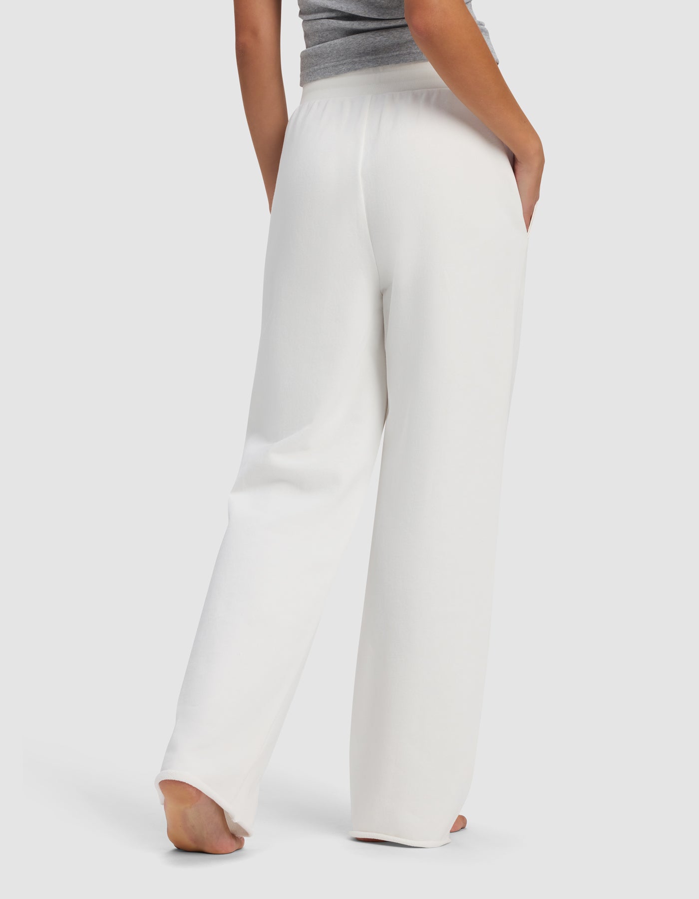 A woman stands barefoot, facing away, wearing Cozy Earth's Women's Chelsea Pant in white with a gray top. One hand is in her pocket, set against a plain white background.