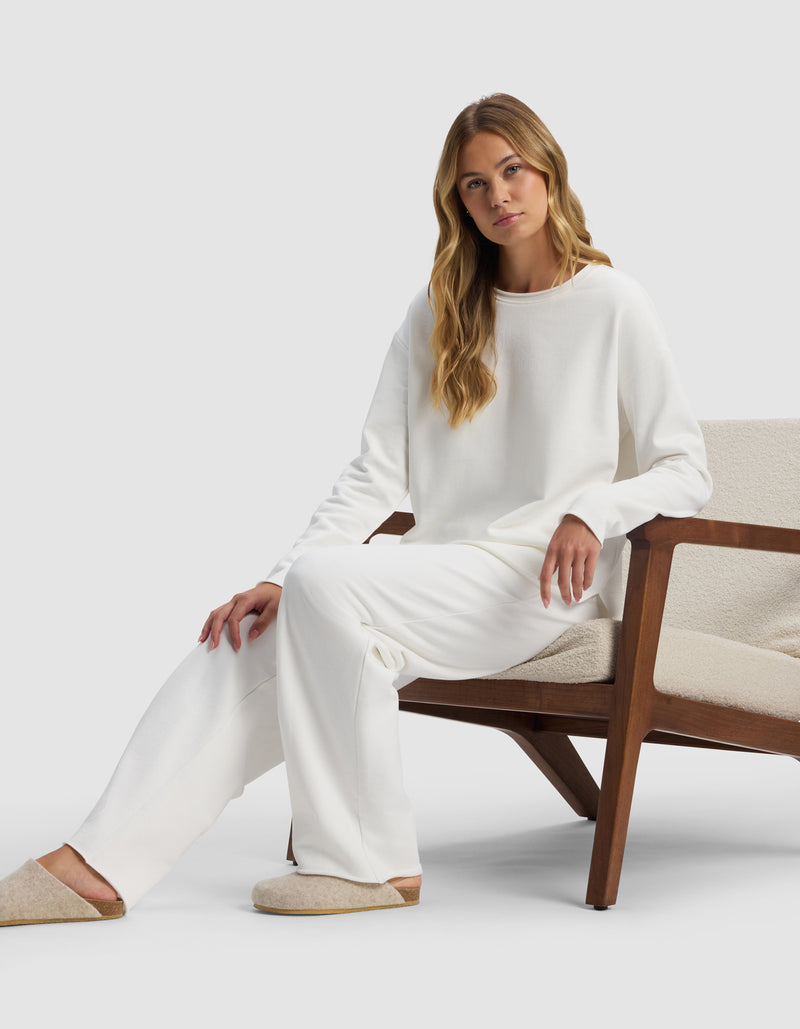 A woman with long blonde hair sits on a wooden chair with cream cushions, wearing Cozy Earth's Women's Chelsea Pant, a loose-fitting white sweater, and beige slippers, against a plain light background.