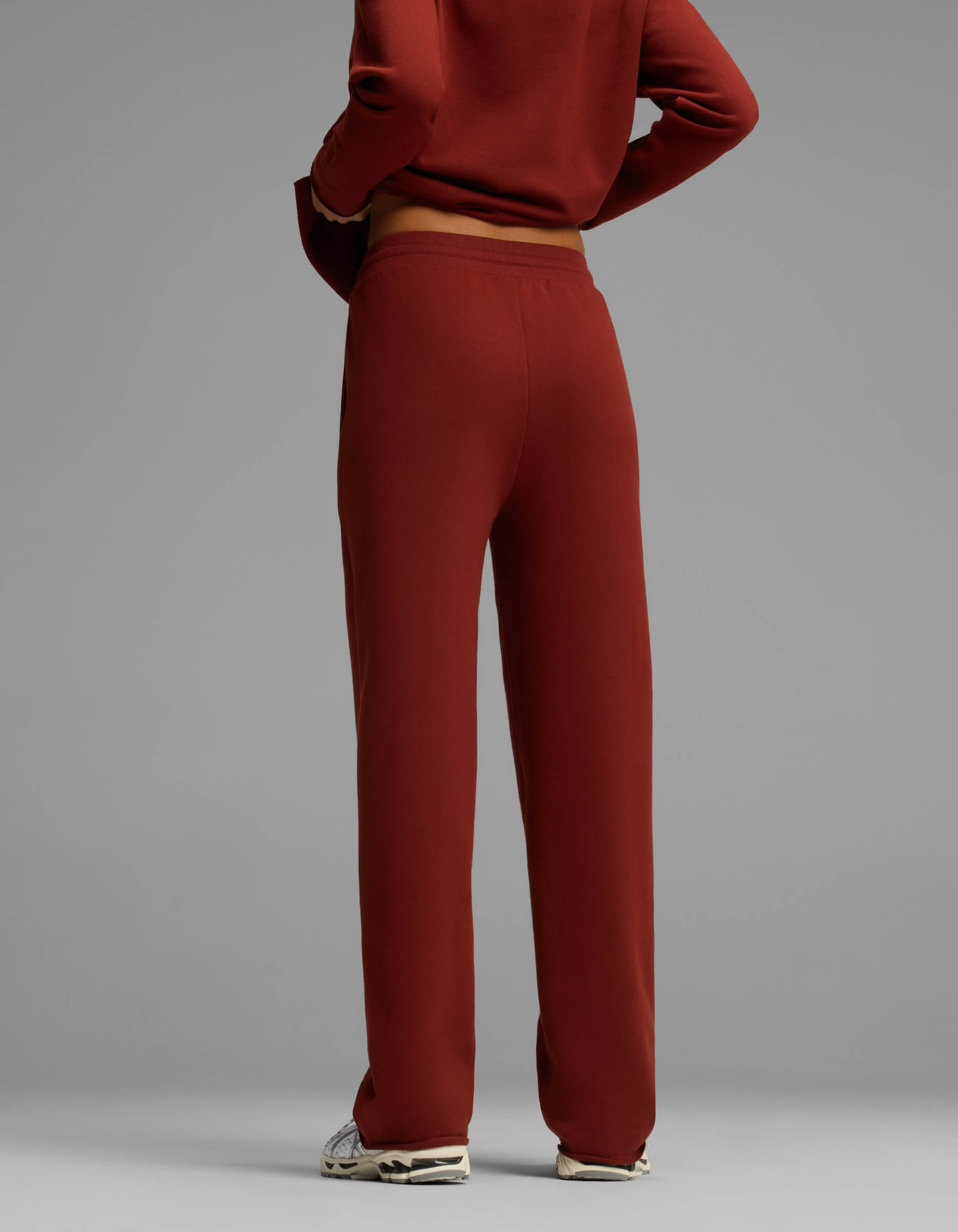 A woman is shown from the back wearing Cozy Earth's Women's Chelsea Pant in a red-brown hue, paired with a matching top. The image focuses on her lower torso and legs, with arms crossed, set against a plain gray background.