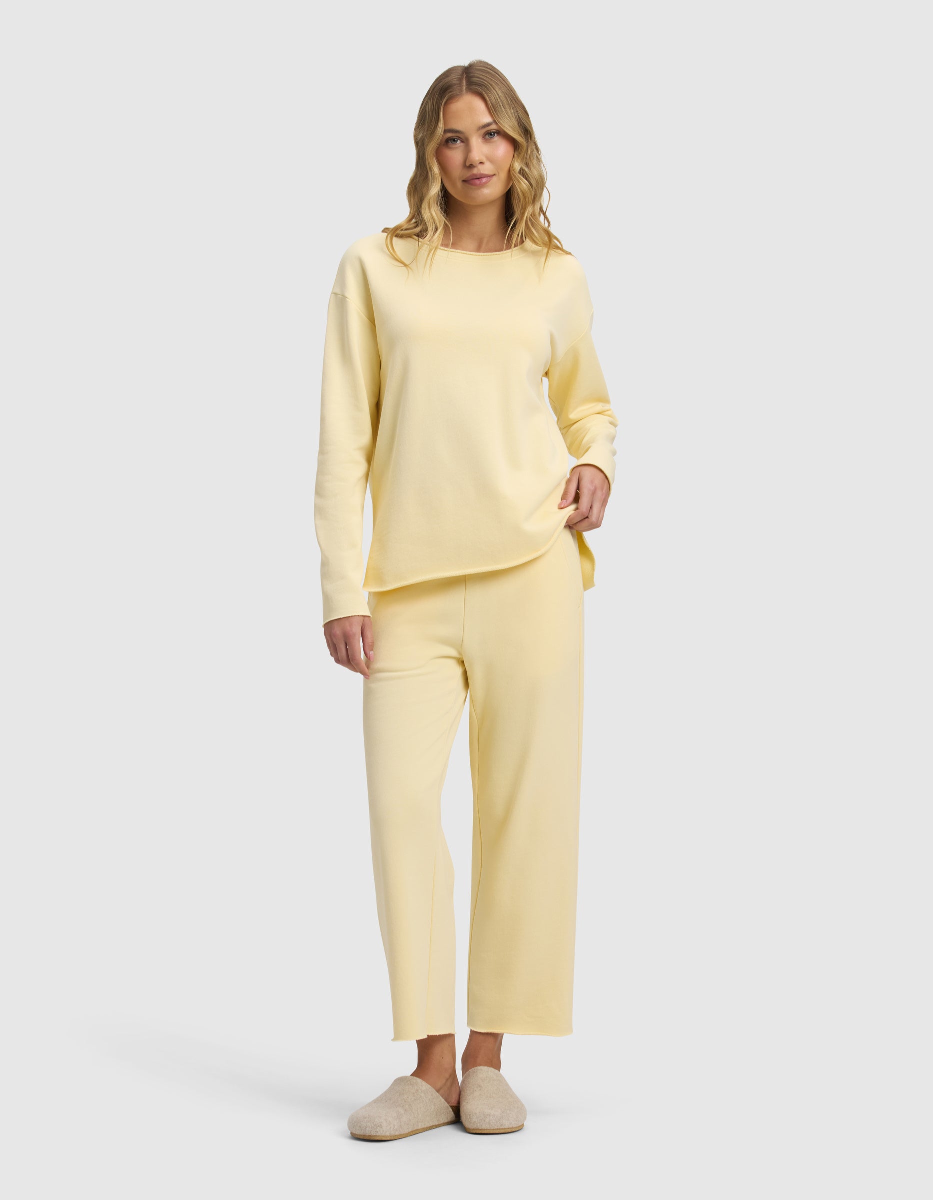 A smiling woman with blonde wavy hair is standing against a light gray background, wearing Cozy Earth's Women's Chelsea Cropped Set in light yellow and beige slippers.