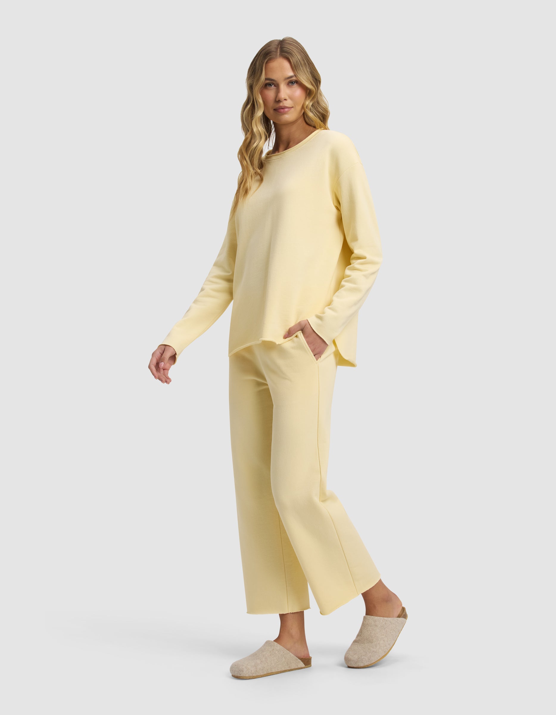 A woman with long wavy hair models the Cozy Earth Women's Chelsea Cropped Set in light yellow, featuring long sleeves and wide-leg pants. She stands sideways with one hand in her pocket, wearing beige slippers against a plain light gray background.|Color:Meringue