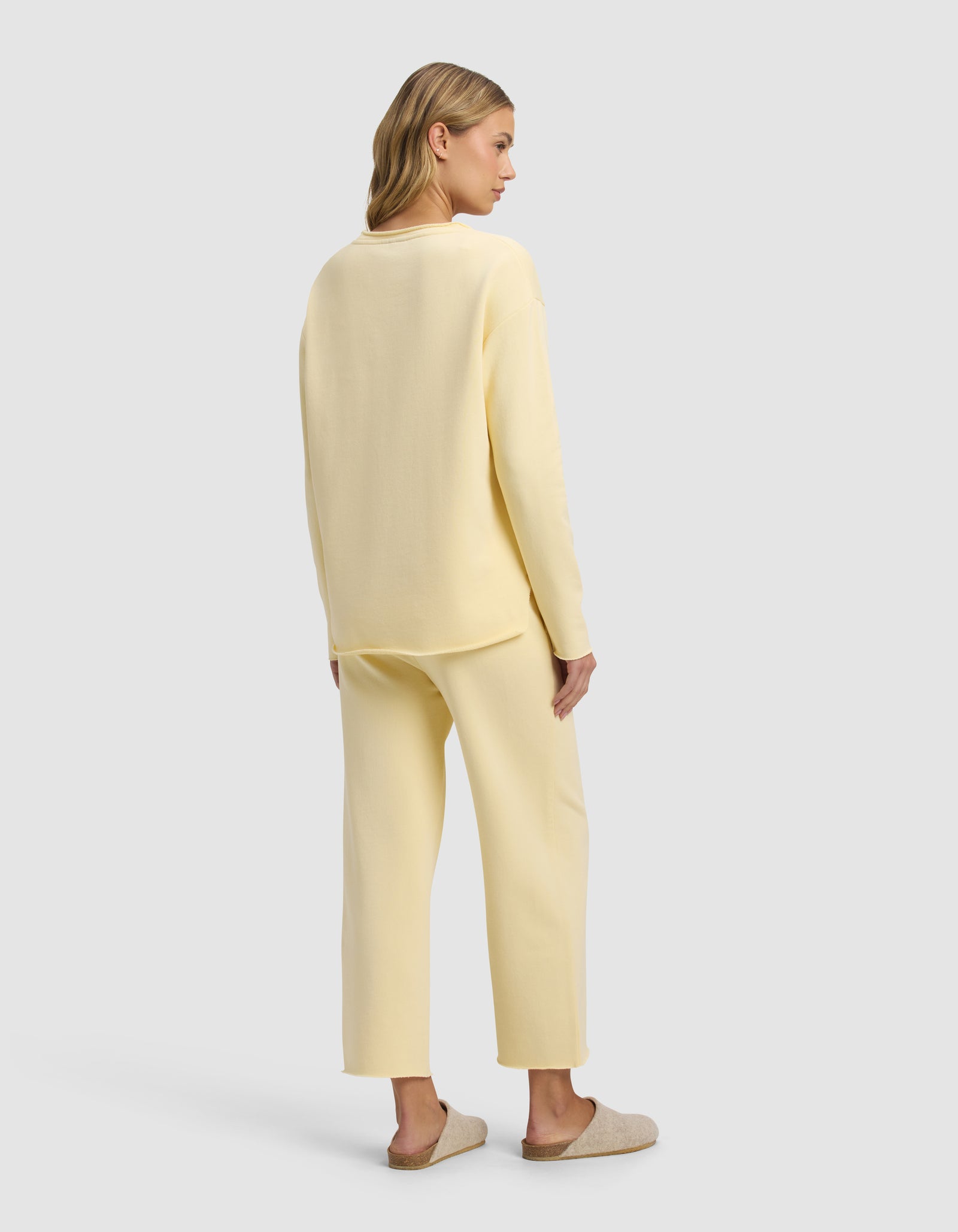 A woman with light brown hair stands against a plain light gray background, wearing the Cozy Earth Women's Chelsea Cropped Set in pale yellow with tan slippers. The set features a loose, long-sleeve top and matching pants.
