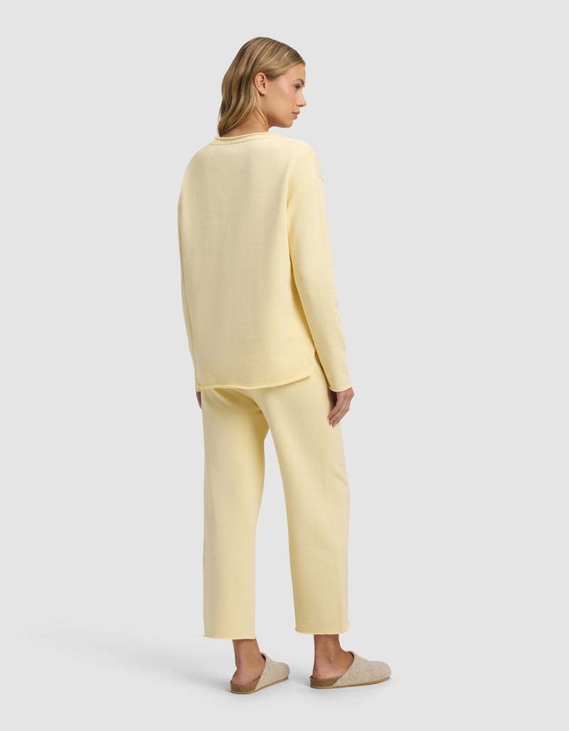 A woman with light brown hair stands against a plain light gray background, wearing the Cozy Earth Women's Chelsea Cropped Set in pale yellow with tan slippers. The set features a loose, long-sleeve top and matching pants.