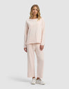 A woman with wavy blonde hair and a neutral expression models the Cozy Earth Women's Chelsea Cropped Set—loose-fitting, light pink long-sleeve top and matching wide-leg pants—paired with white sneakers against a plain background.|Color:Petal