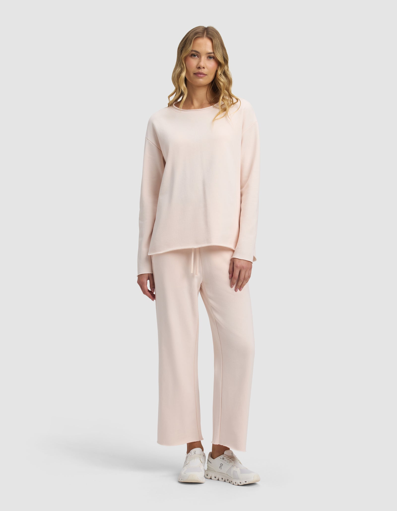 A woman with wavy blonde hair and a neutral expression models the Cozy Earth Women's Chelsea Cropped Set—loose-fitting, light pink long-sleeve top and matching wide-leg pants—paired with white sneakers against a plain background.