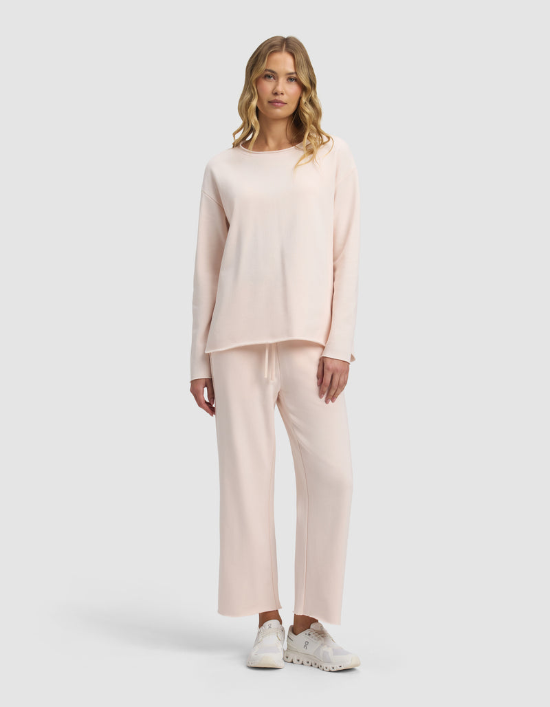 A woman with wavy blonde hair and a neutral expression models the Cozy Earth Women's Chelsea Cropped Set—loose-fitting, light pink long-sleeve top and matching wide-leg pants—paired with white sneakers against a plain background.