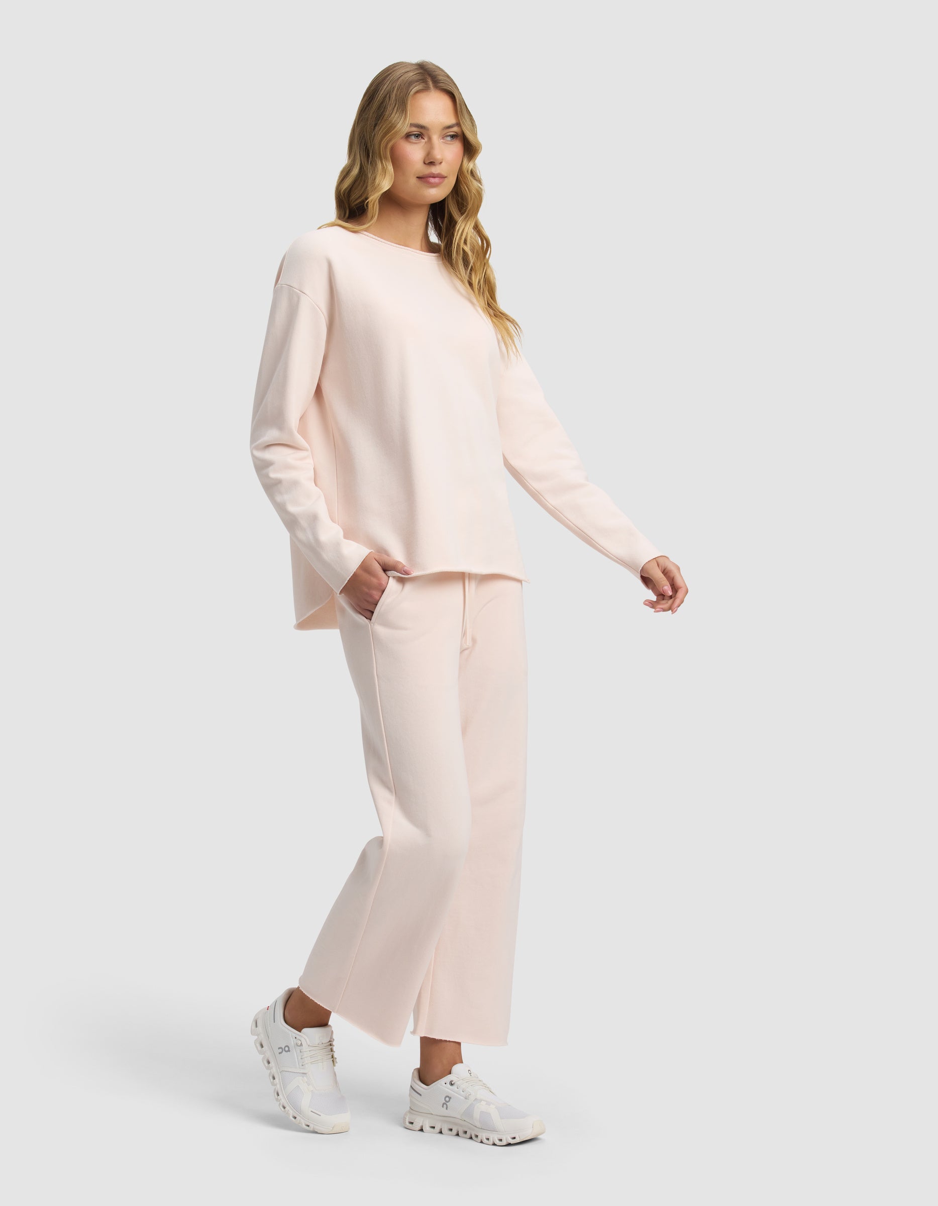 A woman with long blonde hair wears the Cozy Earth Women's Chelsea Cropped Set in light pink, paired with white sneakers, standing against a plain light gray background.|Color:Petal