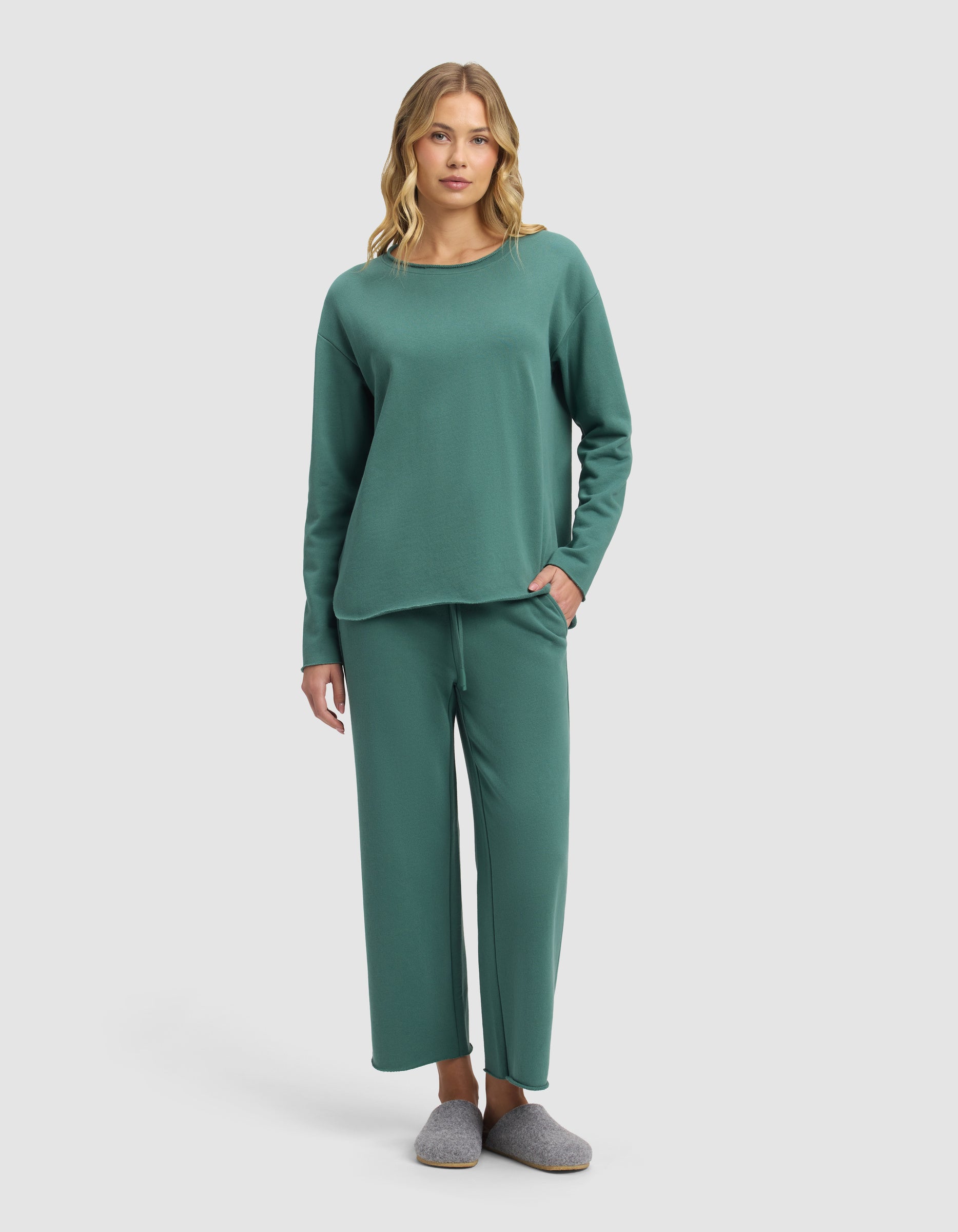 A woman with wavy blond hair stands against a plain background wearing the Cozy Earth Women's Chelsea Cropped Set in green, featuring a matching long-sleeve top and loose pants. She has her left hand in her pocket and is also wearing gray slippers.