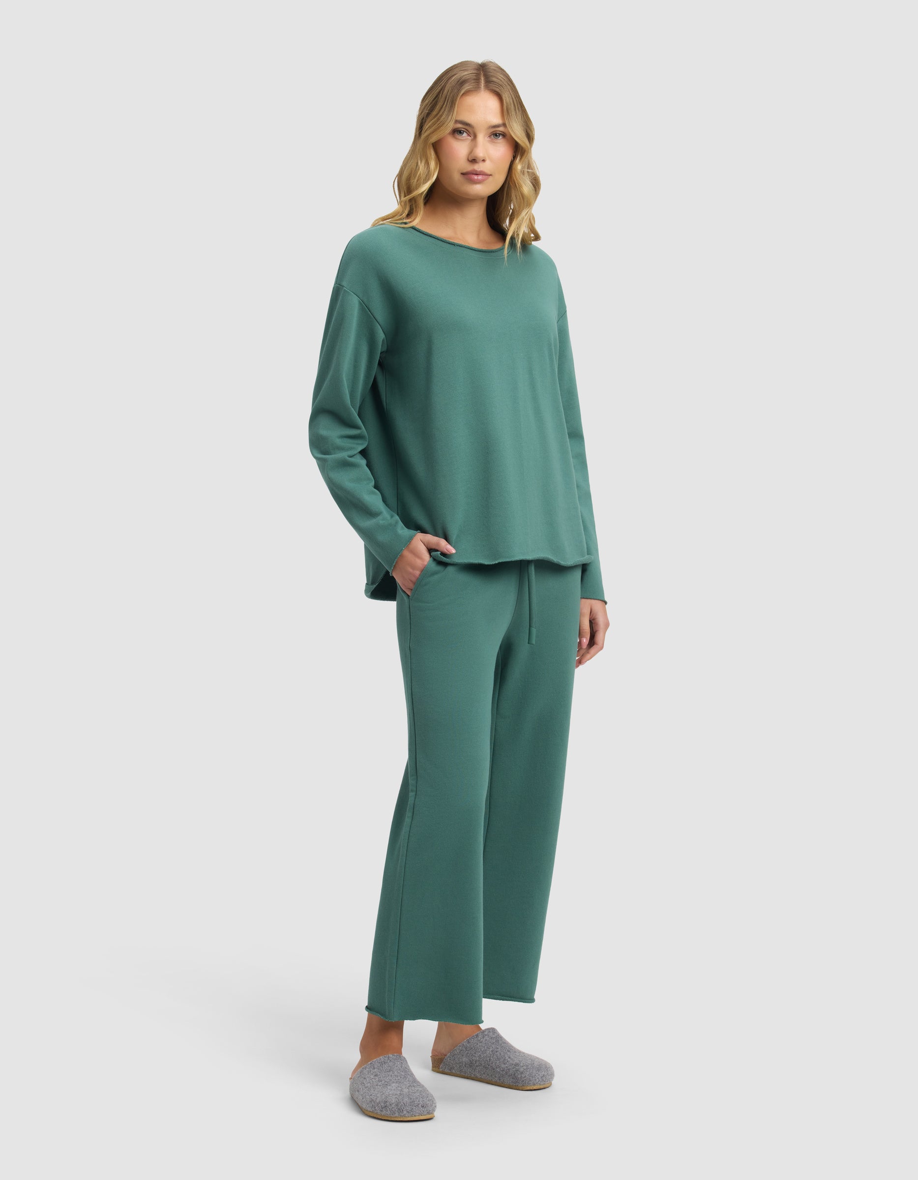 A woman with blonde hair stands against a plain background wearing the Cozy Earth Women's Chelsea Cropped Set in green—featuring a loose, long-sleeve top and wide-leg pants—with gray slippers and her hands in her pockets.