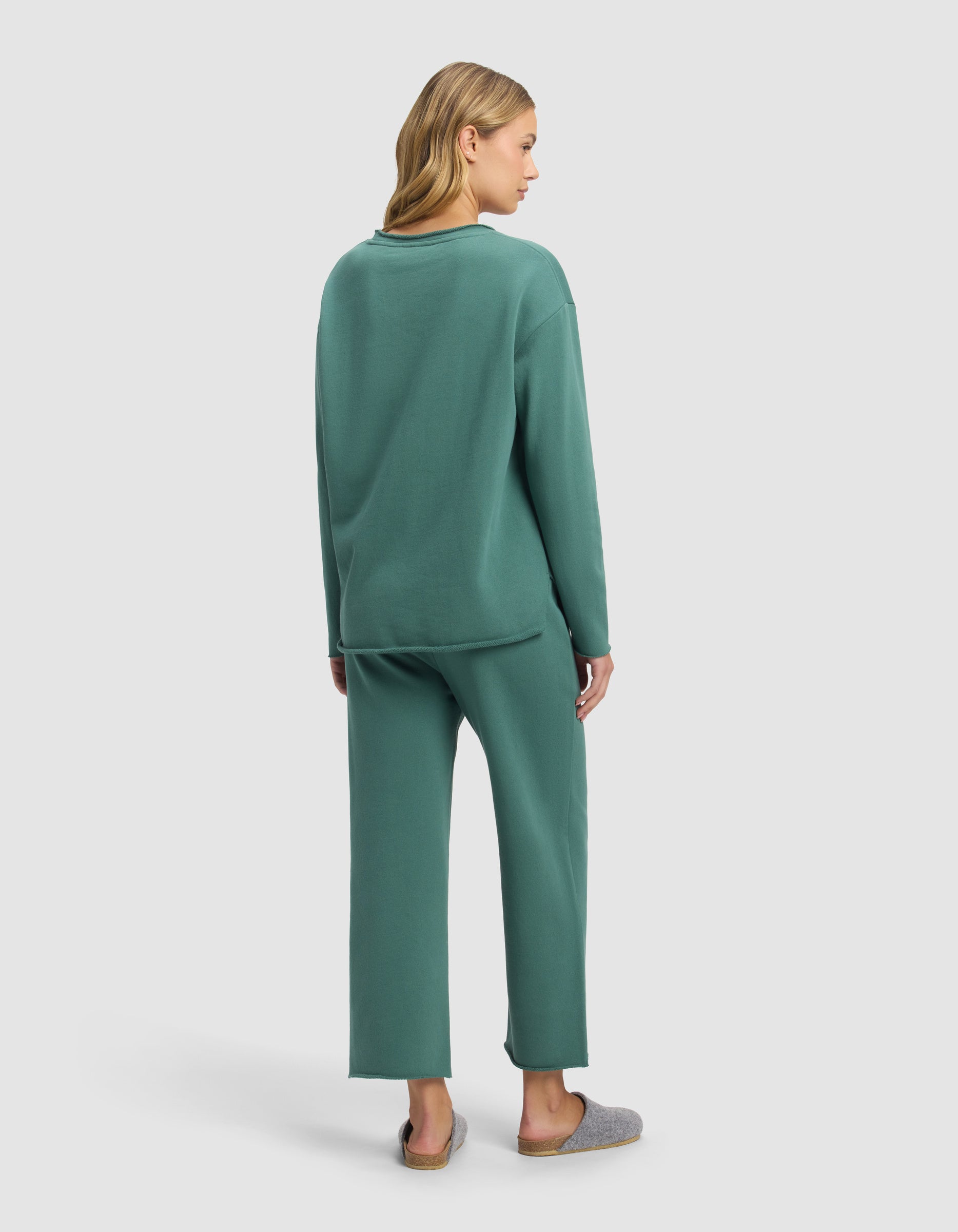 A woman with blonde hair, seen from behind, wears the Cozy Earth Women's Chelsea Cropped Set in green with gray slippers, standing against a plain light background.