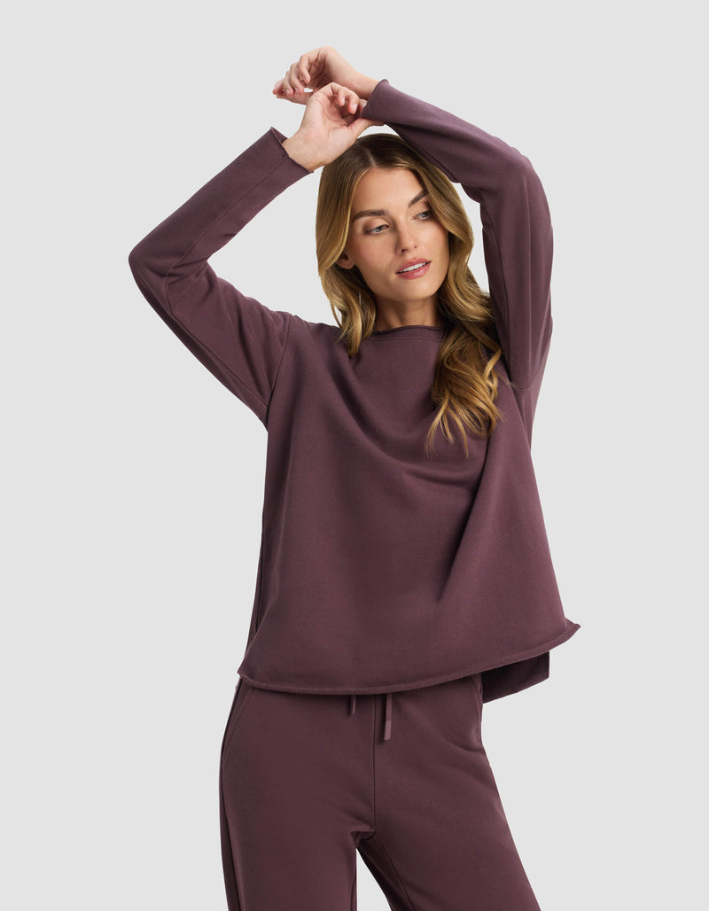A woman models the Cozy Earth Women's Chelsea Pullover in mauve, paired with matching pants, standing against a light gray background with her arms raised and crossed above her head, gazing to the side.