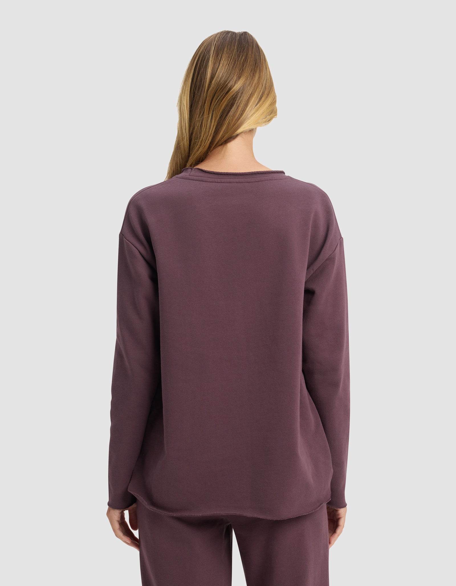 A person with long, light brown hair faces away, wearing the Cozy Earth Women's Chelsea Pullover and matching pants in mauve against a plain light gray background.