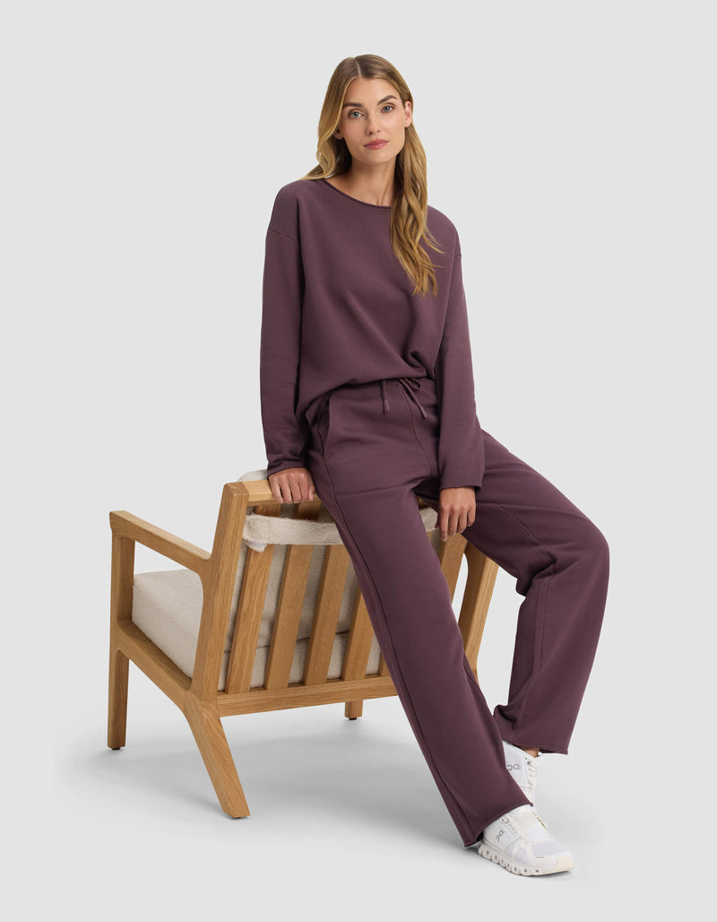 A woman wearing Cozy Earth's Women's Chelsea Pullover in dark mauve and matching pants sits on the armrest of a wooden chair with beige cushions, her long hair down and white sneakers on, looking calmly at the camera.