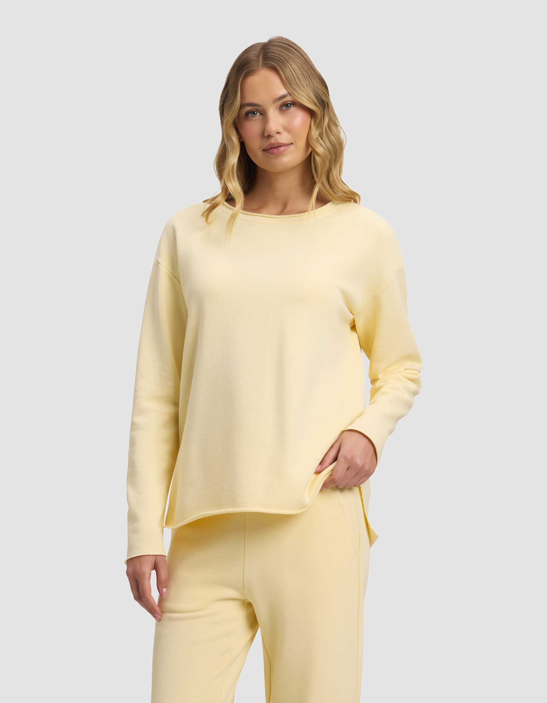 A woman with blonde wavy hair models the Cozy Earth Women's Chelsea Pullover, a loose-fitting pale yellow long-sleeve top with matching pants, standing against a plain light gray background.