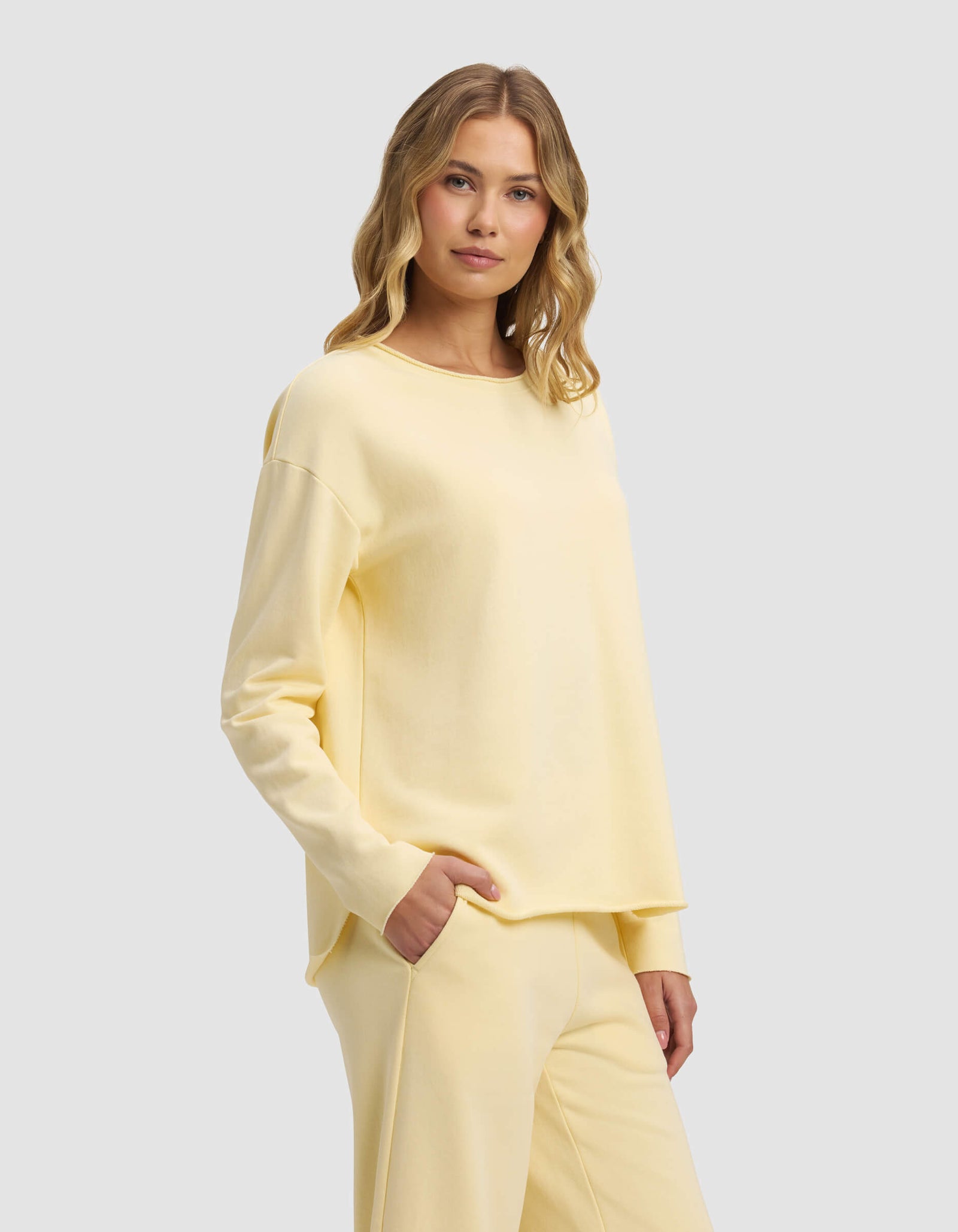 A woman with wavy blonde hair models the Cozy Earth Women's Chelsea Pullover and matching pants in pale yellow, standing against a light gray background with one hand in her pocket.