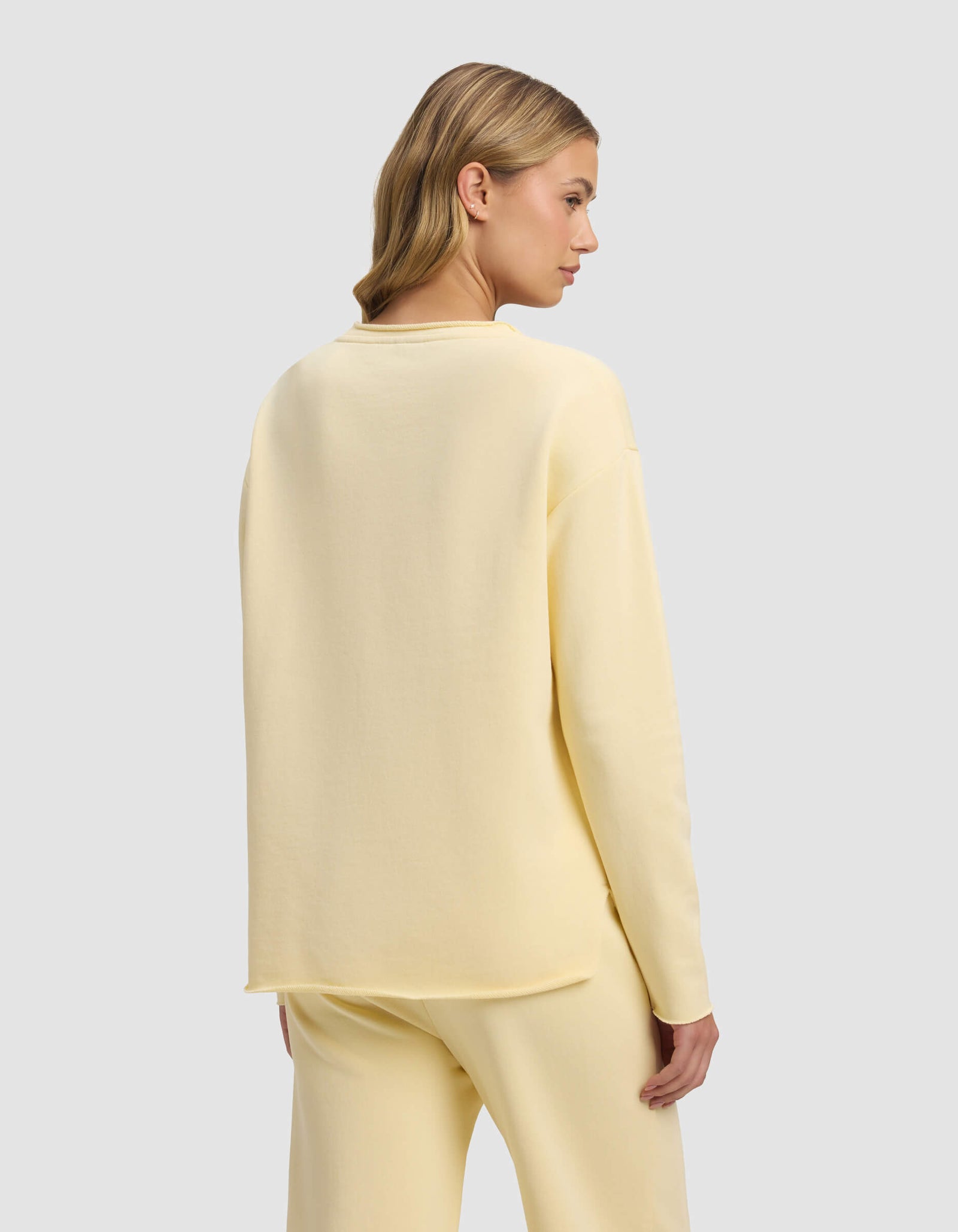 A woman with blonde hair styled in loose waves wears the Cozy Earth Women's Chelsea Pullover in pale yellow, paired with matching pants. She stands turned slightly away, displaying the back of her outfit against a light gray background.