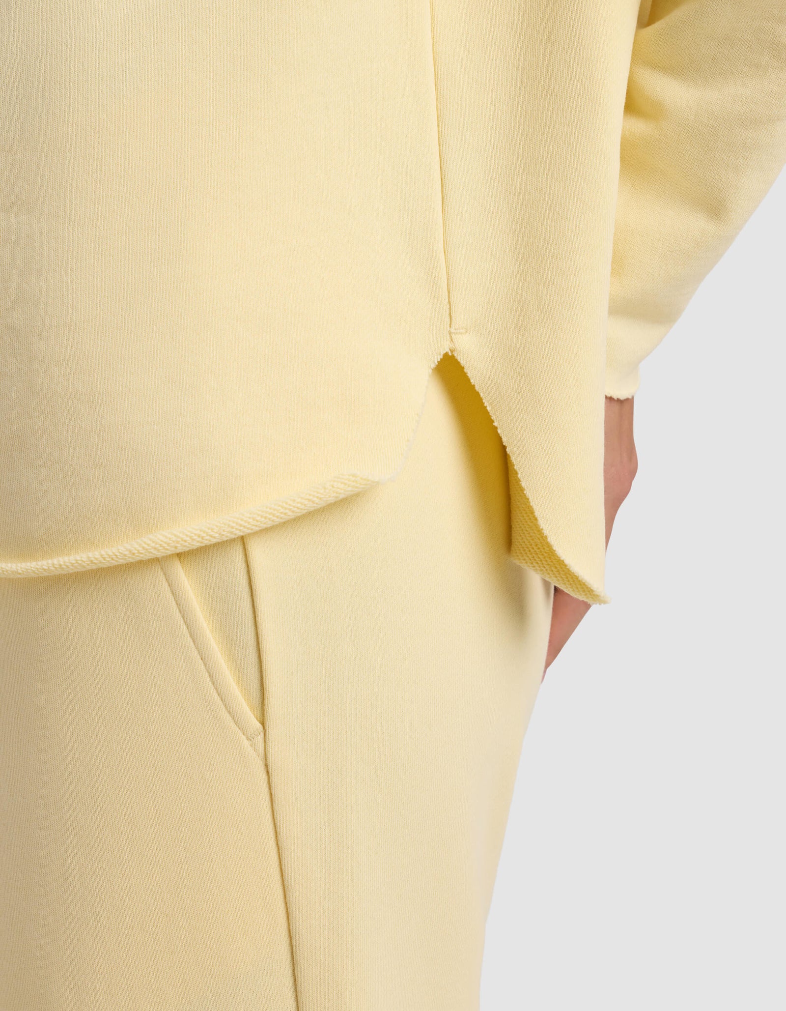 A close-up of a person wearing the Cozy Earth Women's Chelsea Pullover in pale yellow, featuring soft knit fabric, a curved hem with a side slit, and matching pants. Their hand is in the pocket against a plain light gray background.