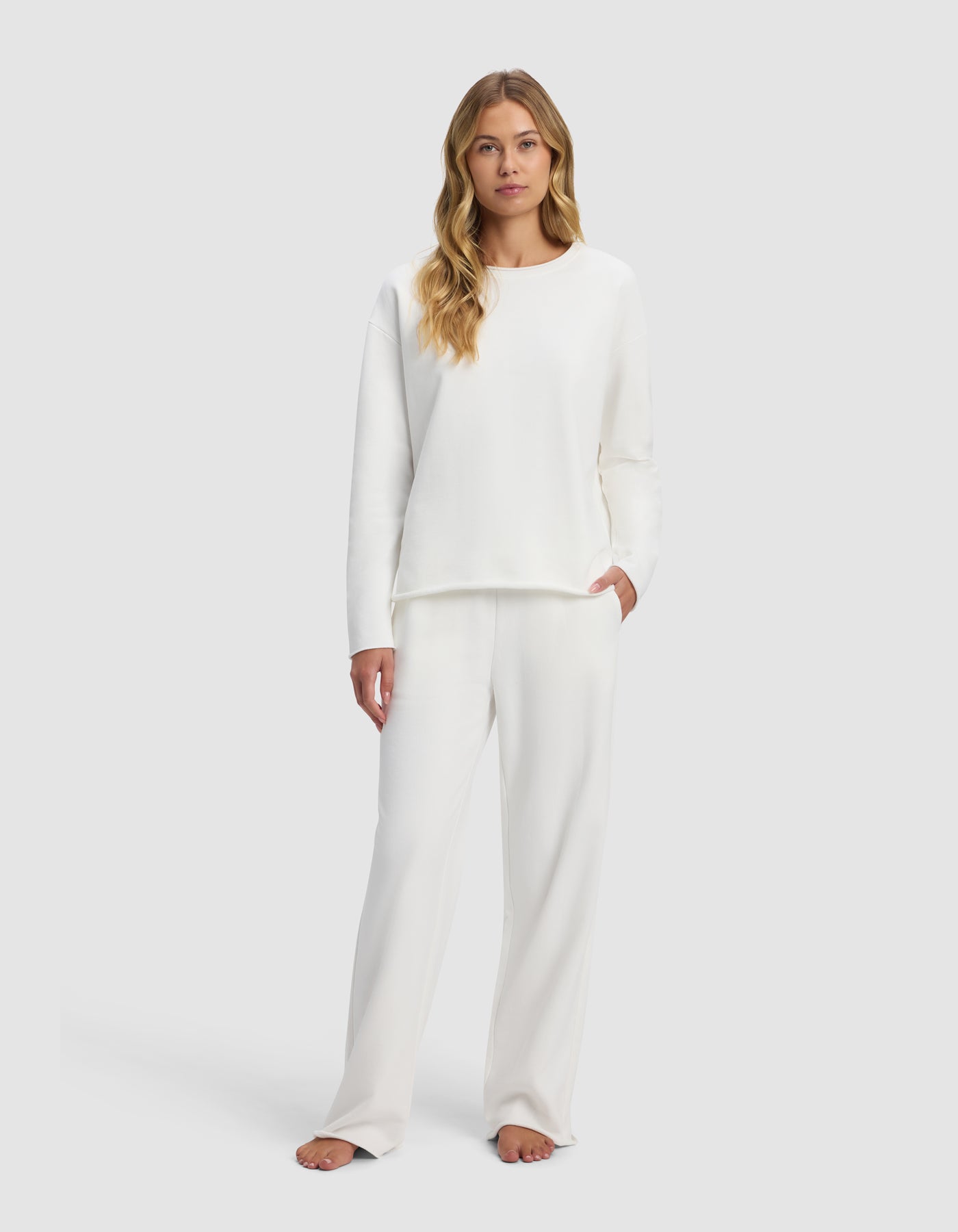 A woman with long blonde hair models the Cozy Earth Women's Chelsea Set—a loose-fitting, long-sleeve white top and matching pants—standing barefoot against a light gray background, gazing at the camera with a neutral expression.