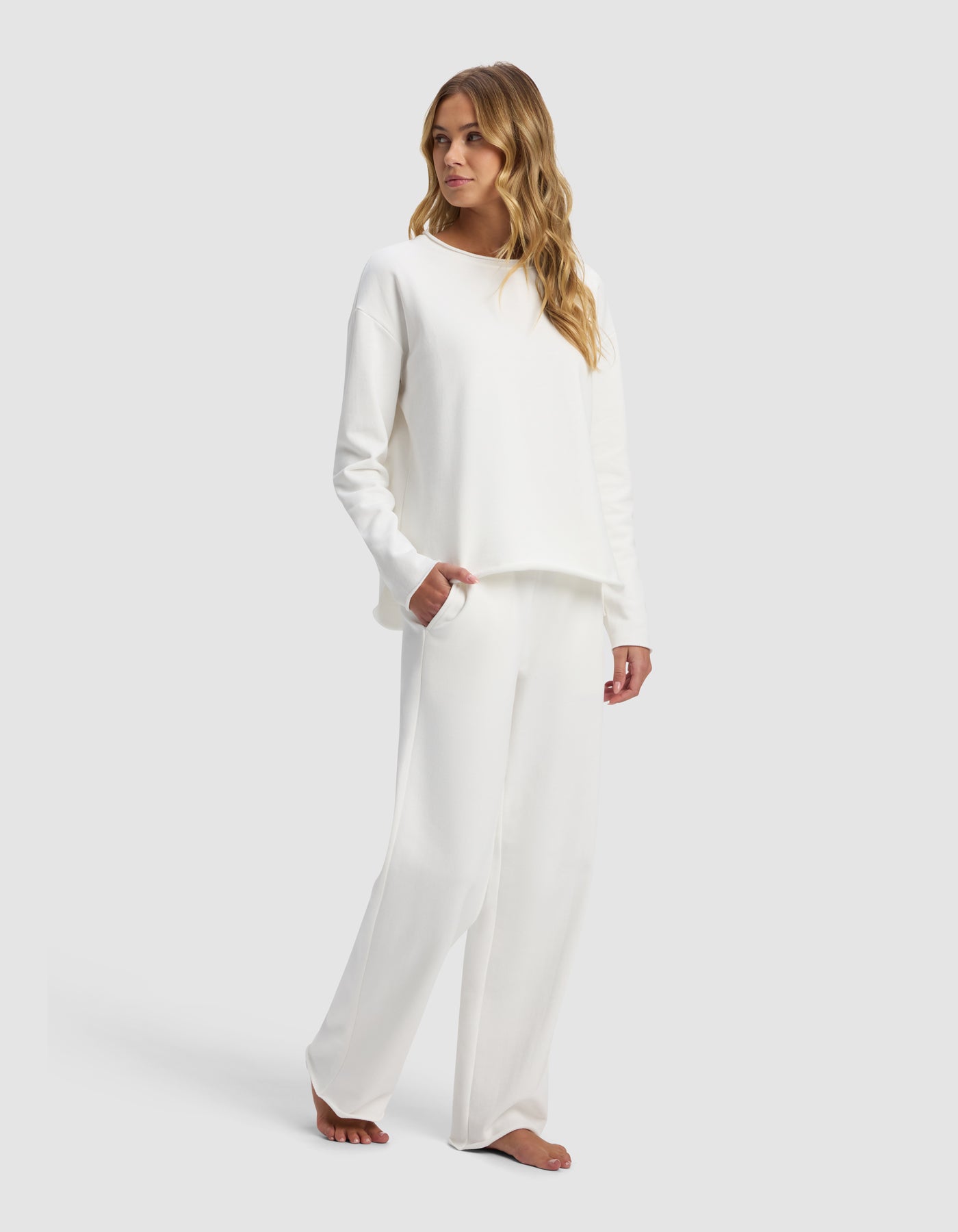 A woman with long wavy hair stands barefoot against a plain light background, wearing the Women's Chelsea Set by Cozy Earth—a loose-fitting, long-sleeve white top and matching wide-leg white pants.|Color:Bone