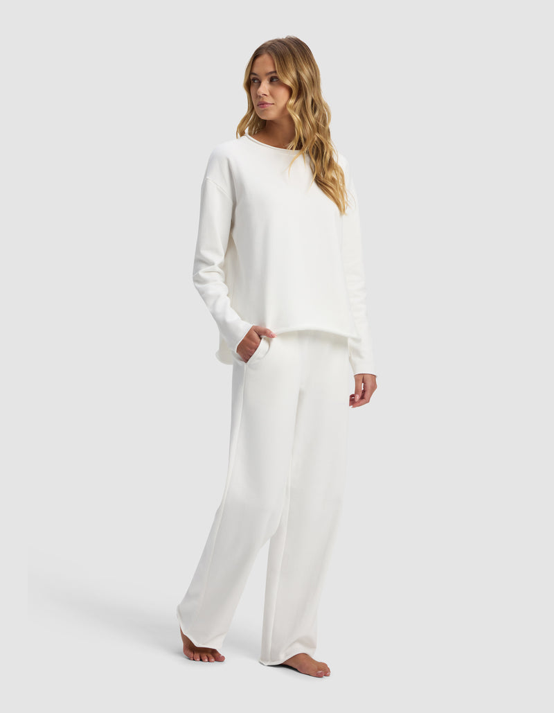 A woman with long wavy hair stands barefoot against a plain light background, wearing the Women's Chelsea Set by Cozy Earth—a loose-fitting, long-sleeve white top and matching wide-leg white pants.
