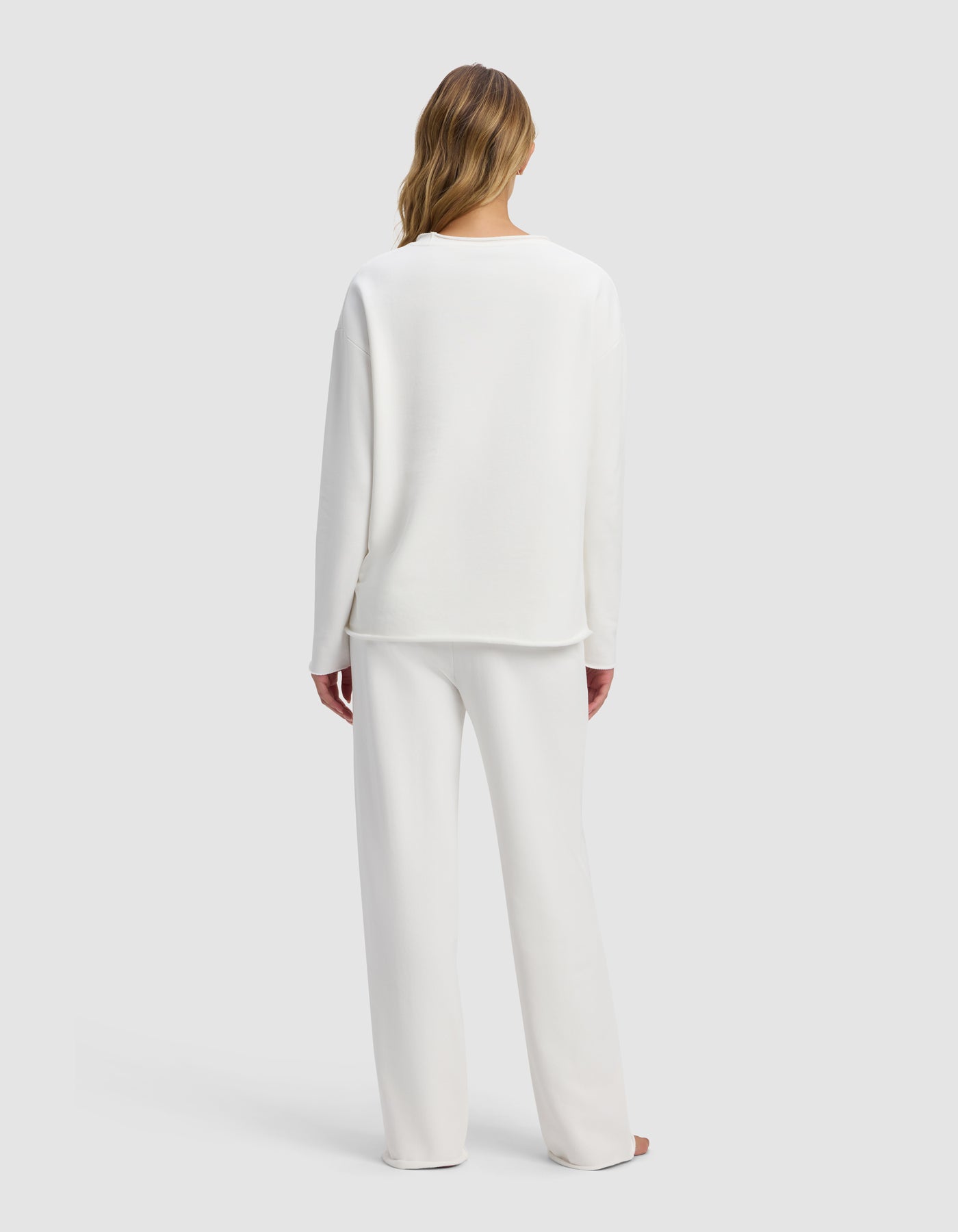 A woman stands barefoot facing away in the Cozy Earth Women's Chelsea Set—loose-fitting white long-sleeve top and pants—against a plain light gray background.
