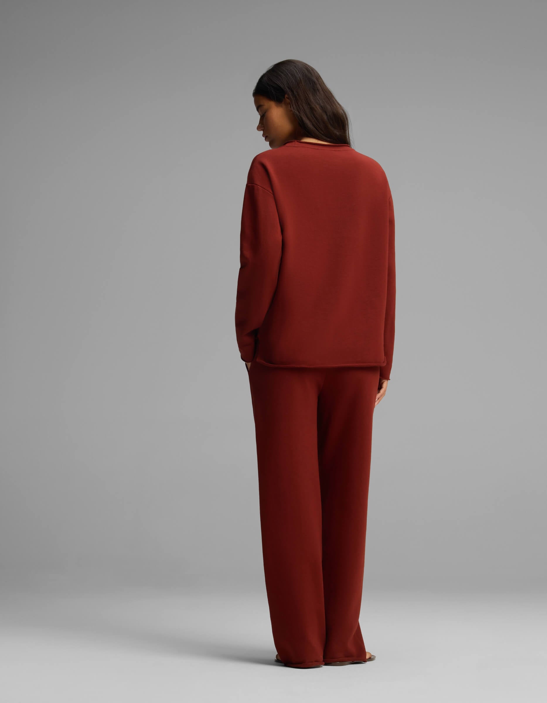 A person with long dark hair stands facing away, wearing the Cozy Earth Women's Chelsea Set—a loose-fitting, long-sleeve dark red top and matching wide-leg pants—against a plain gray background.|Color:Currant