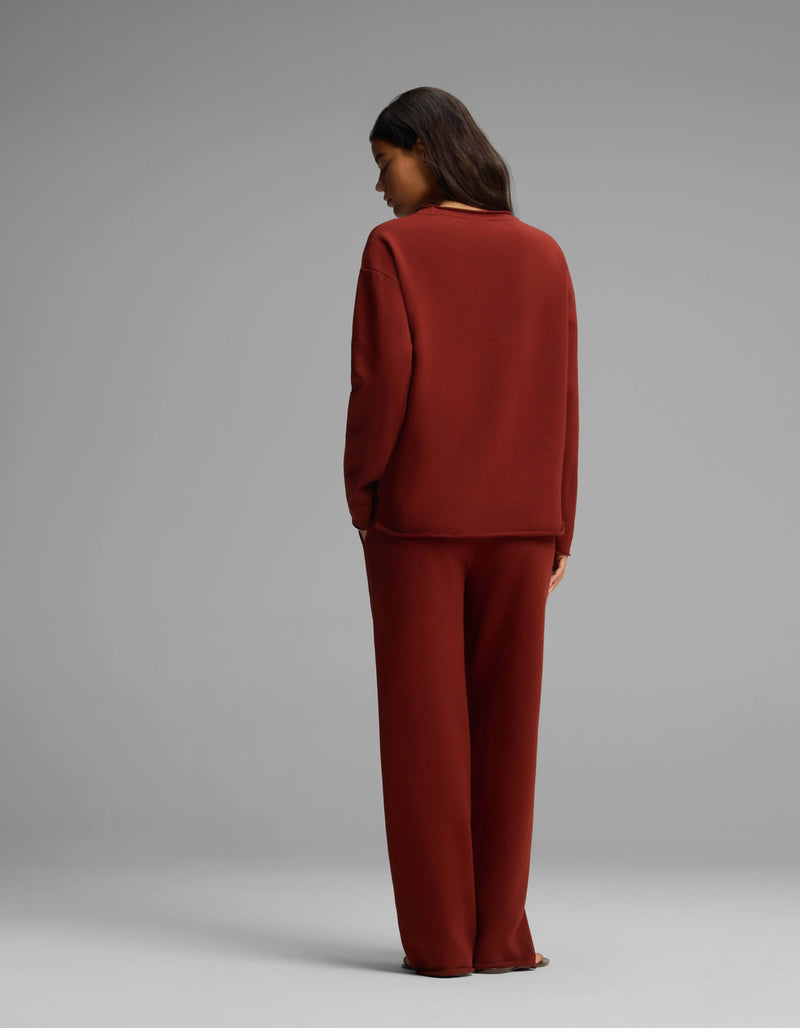 A person with long dark hair stands facing away, wearing the Cozy Earth Women's Chelsea Set—a loose-fitting, long-sleeve dark red top and matching wide-leg pants—against a plain gray background.