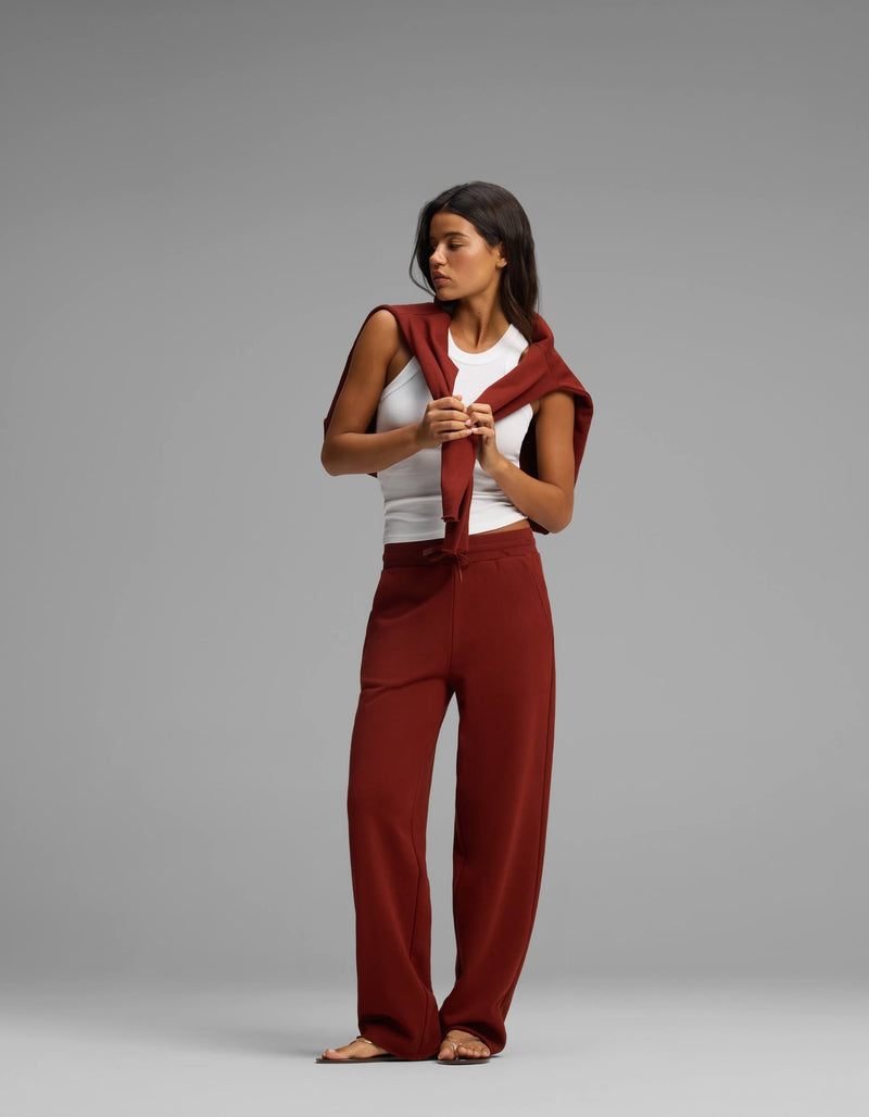 A woman stands against a gray background wearing the Women's Chelsea Set from Cozy Earth, which includes a white tank top, rust-colored pants, and a matching sweater draped over her shoulders. She looks down while holding the sweater’s sleeves in front of her.