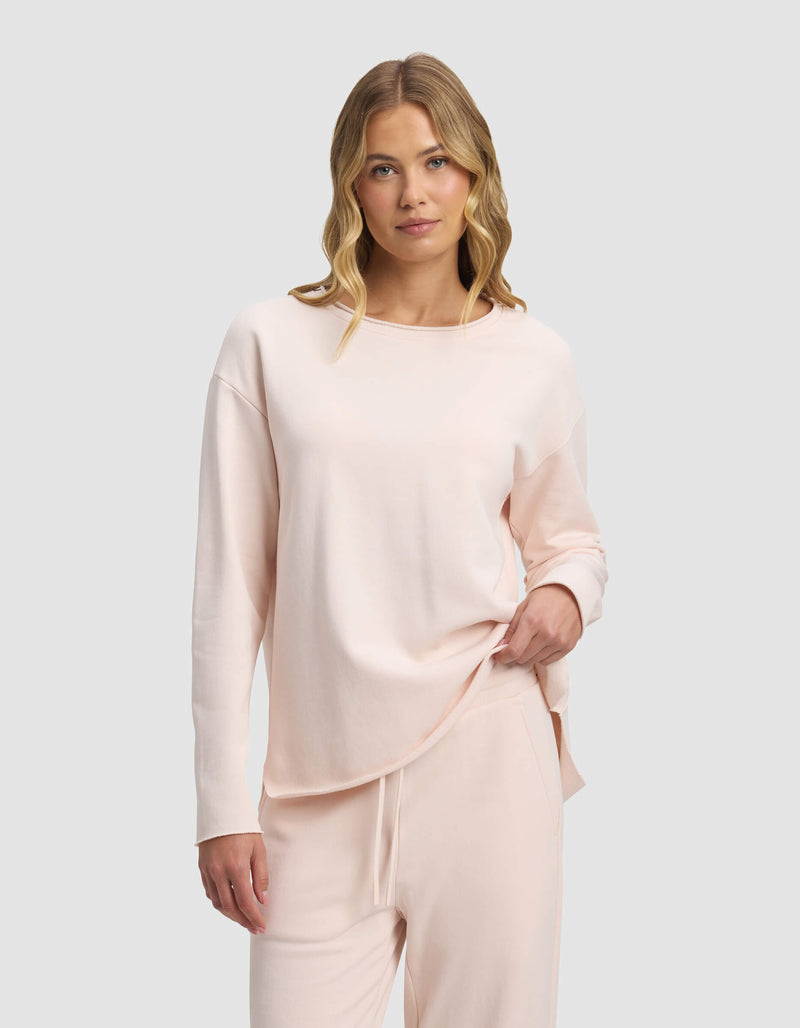 A woman with wavy blonde hair wears the Cozy Earth Women's Chelsea Pullover in light pink and matching drawstring pants, standing against a plain light gray background while looking at the camera and holding part of her shirt hem.